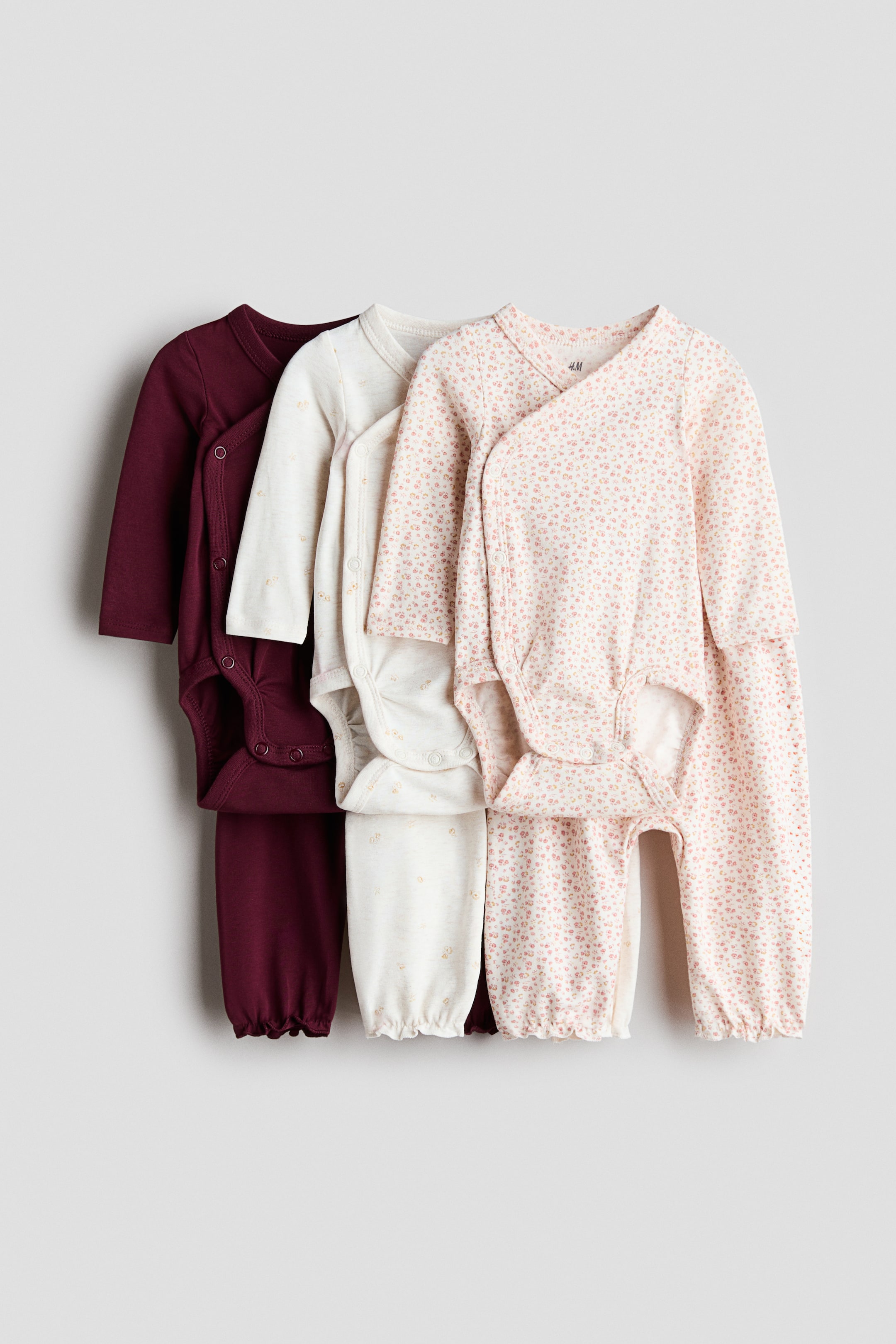 View larger image: Three baby outfits are hung front-facing, with each set featuring a long-sleeve, wrap-front bodysuit and matching pants. The sets are shown in solid burgundy, patterned cream, and pink floral print, with the bodysuits featuring side snap buttons and the pants showing gathered cuffs.