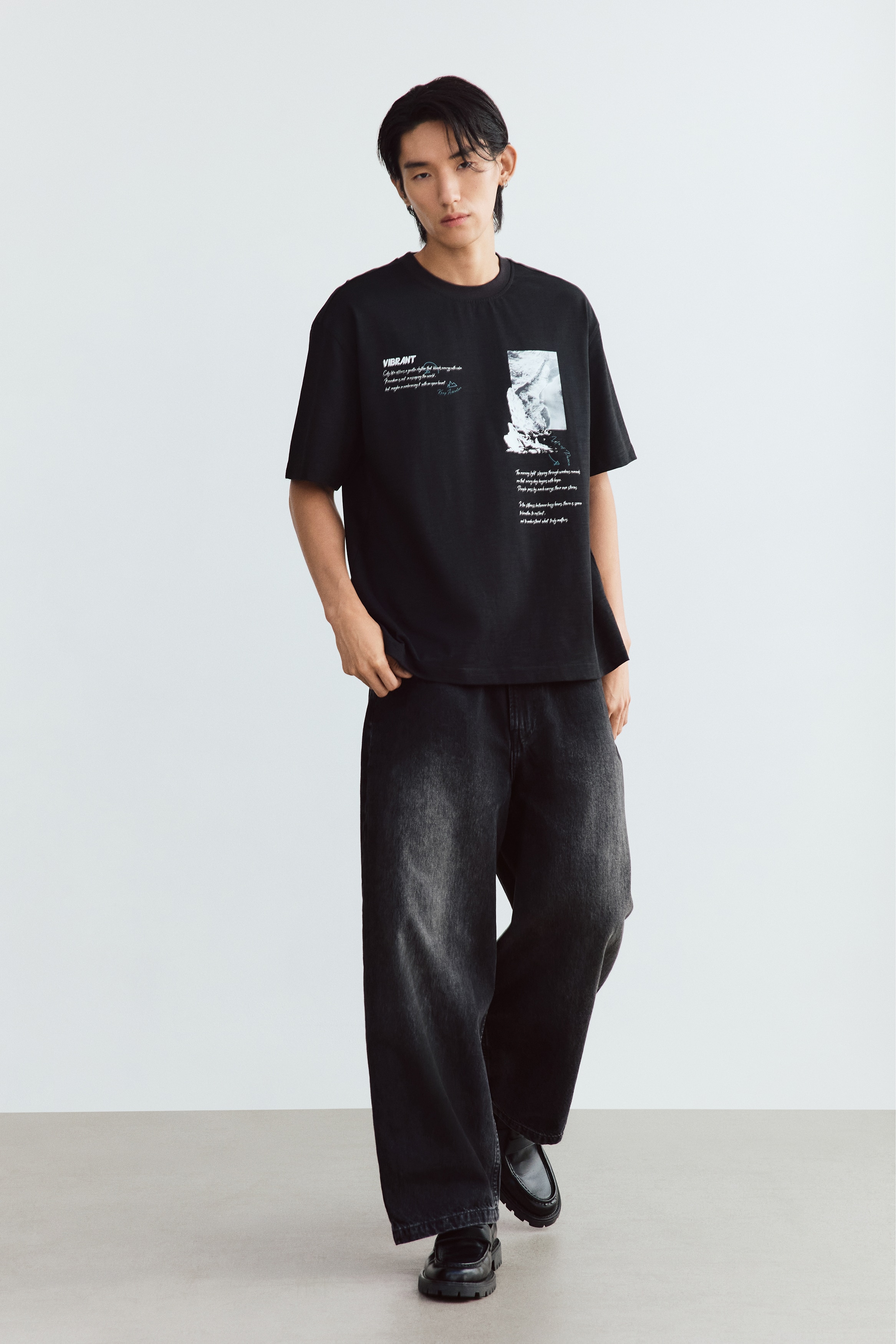 A black heavyweight cotton T-shirt is hanging front-facing, showcasing a loose fit with dropped shoulders, a round ribbed neckline, and a straight hem. The chest features white graphic prints, including the word "VIBRANT" with cursive text on one side and a rectangular landscape graphic with white script below it on the other.