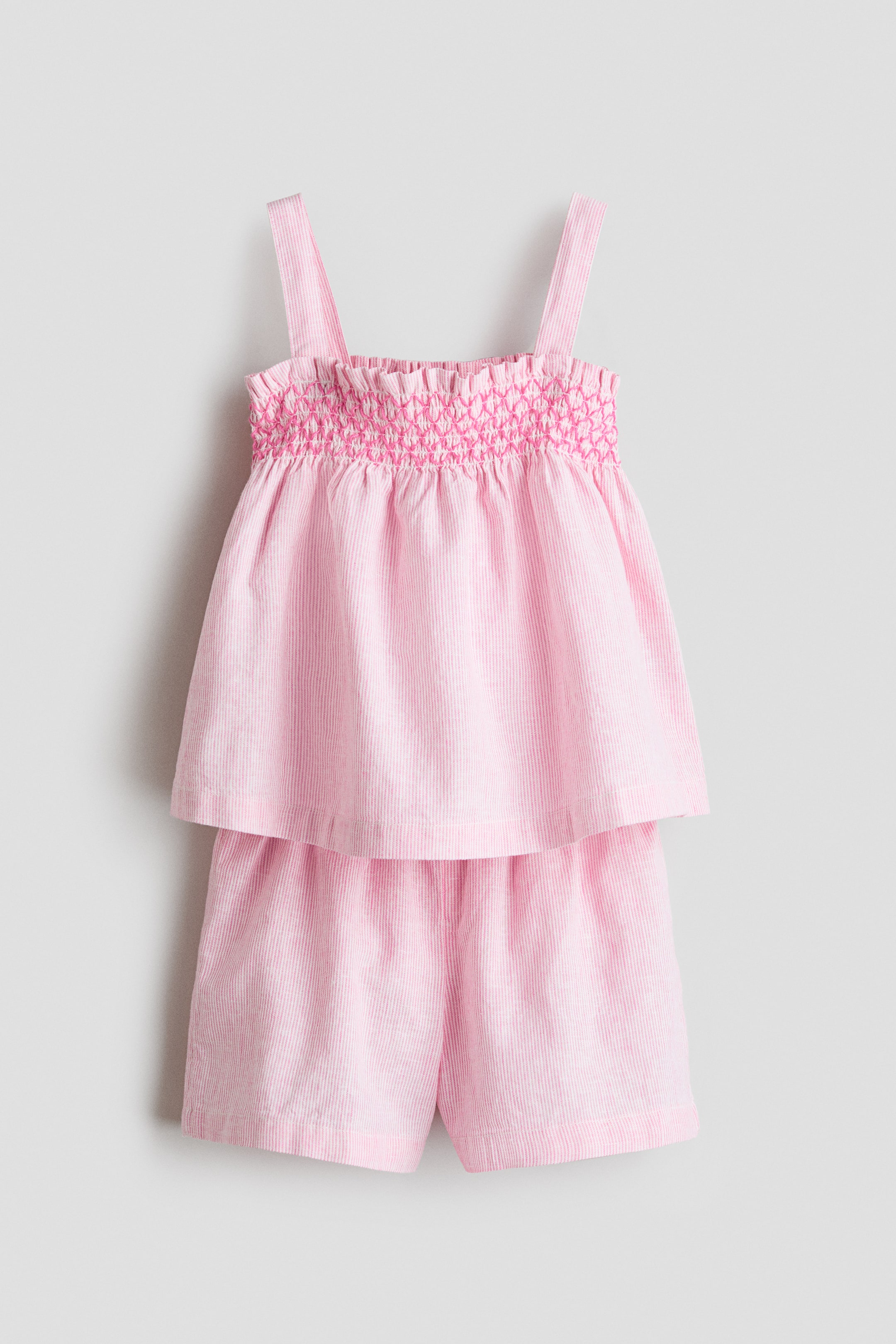 Ver imagen más grande: Un conjunto de rayas rosas y blancas se presenta desde el frente, con un top acampanado de tirantes y pantalones cortos a juego. El top muestra un escote cuadrado con un fruncido rosa oscuro en el pecho, mientras que los pantalones cortos aparecen con un dobladillo simple.