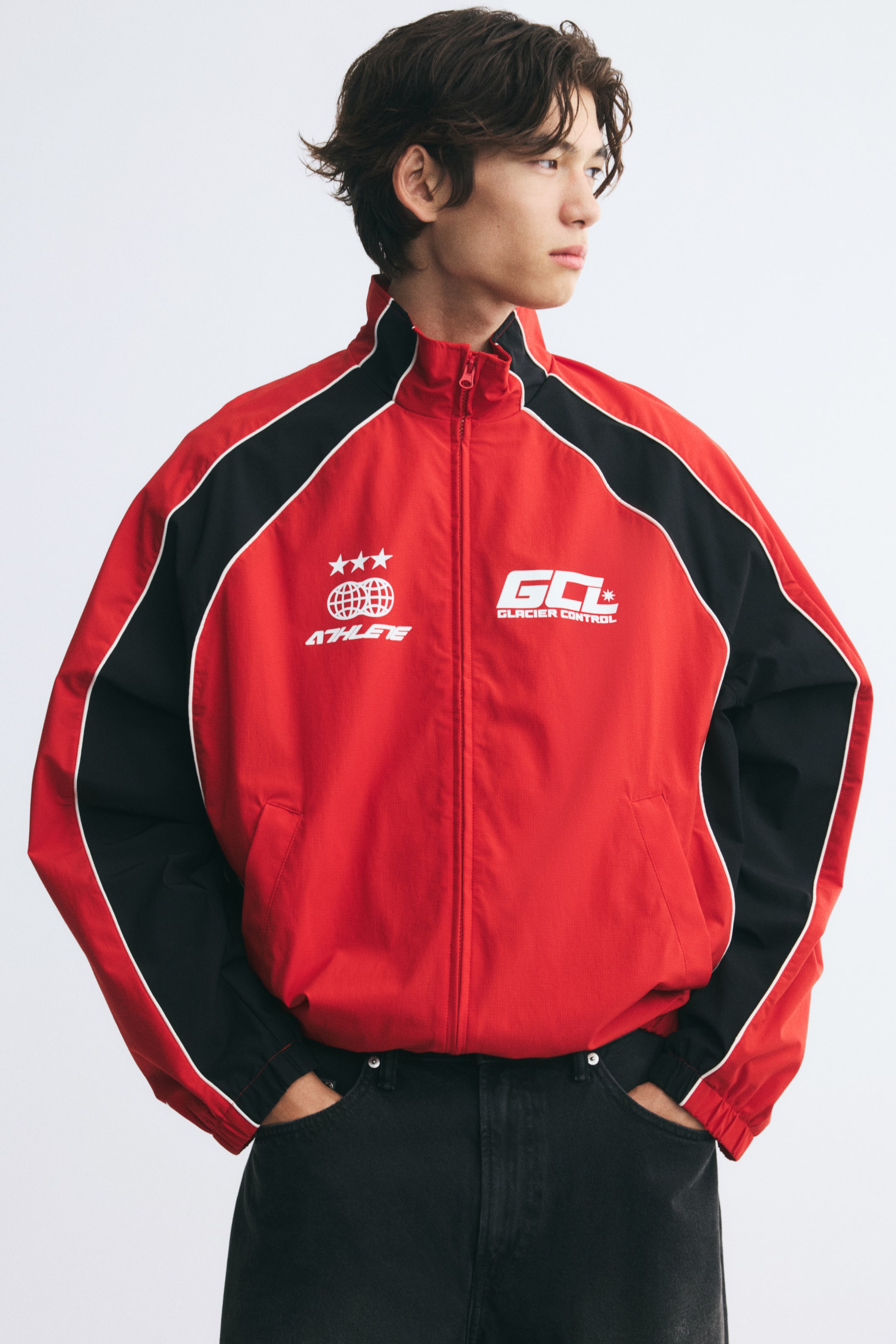 Voir une image plus grande: Une veste de survêtement rouge et noire, avec passepoil blanc et motifs graphiques sur la poitrine, est portée par le mannequin. La veste présente un col montant et des poignets élastiqués, et elle est portée avec un jean noir.