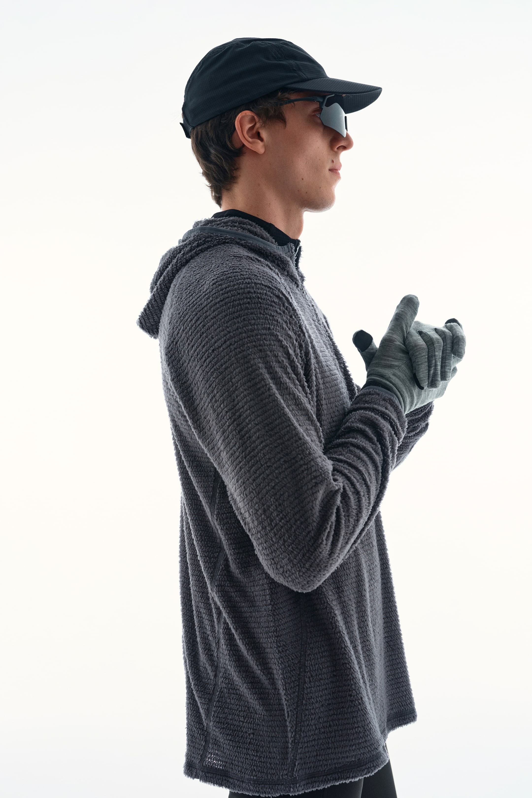 Xem ảnh lớn hơn: Một chiếc áo hoodie màu xám có họa tiết vân nhám với hoa văn ô lưới mịn đang được người mẫu mặc, được thể hiện ở góc nghiêng. Áo hoodie được phối cùng mũ lưỡi trai màu đen, kính râm tối màu và găng tay màu xám.