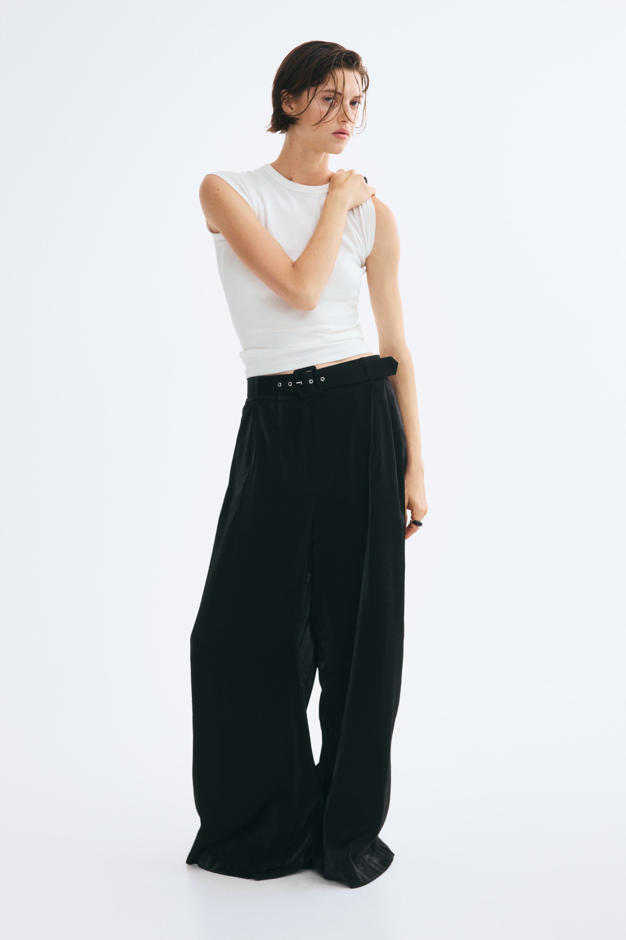 Agrandir l'image: Un pantalon noir à jambes larges, orné de plis sur le devant et d’un léger effet satiné, est porté par le mannequin, associé à une ceinture noire à œillets et à un haut blanc sans manches. Ce pantalon taille haute à la coupe ample tombe jusqu’au sol.