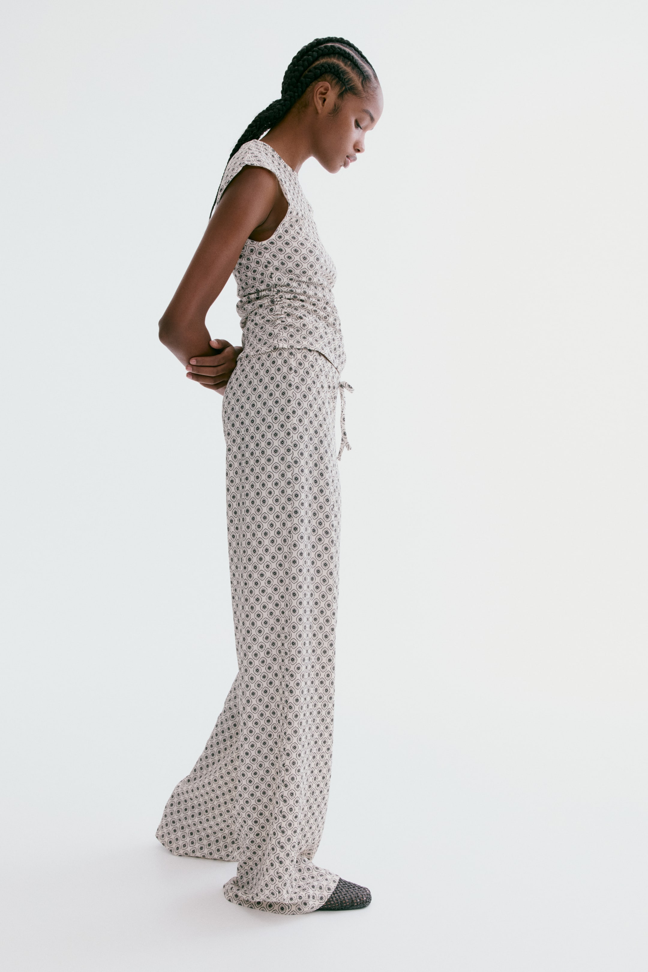 View larger image: A two-piece outfit, featuring a cap-sleeve top and wide-leg trousers, is shown from a side view. Both pieces are patterned with repeating light beige and dark grey concentric ovals. The top gathers at the waist, and the trousers include a visible drawstring.