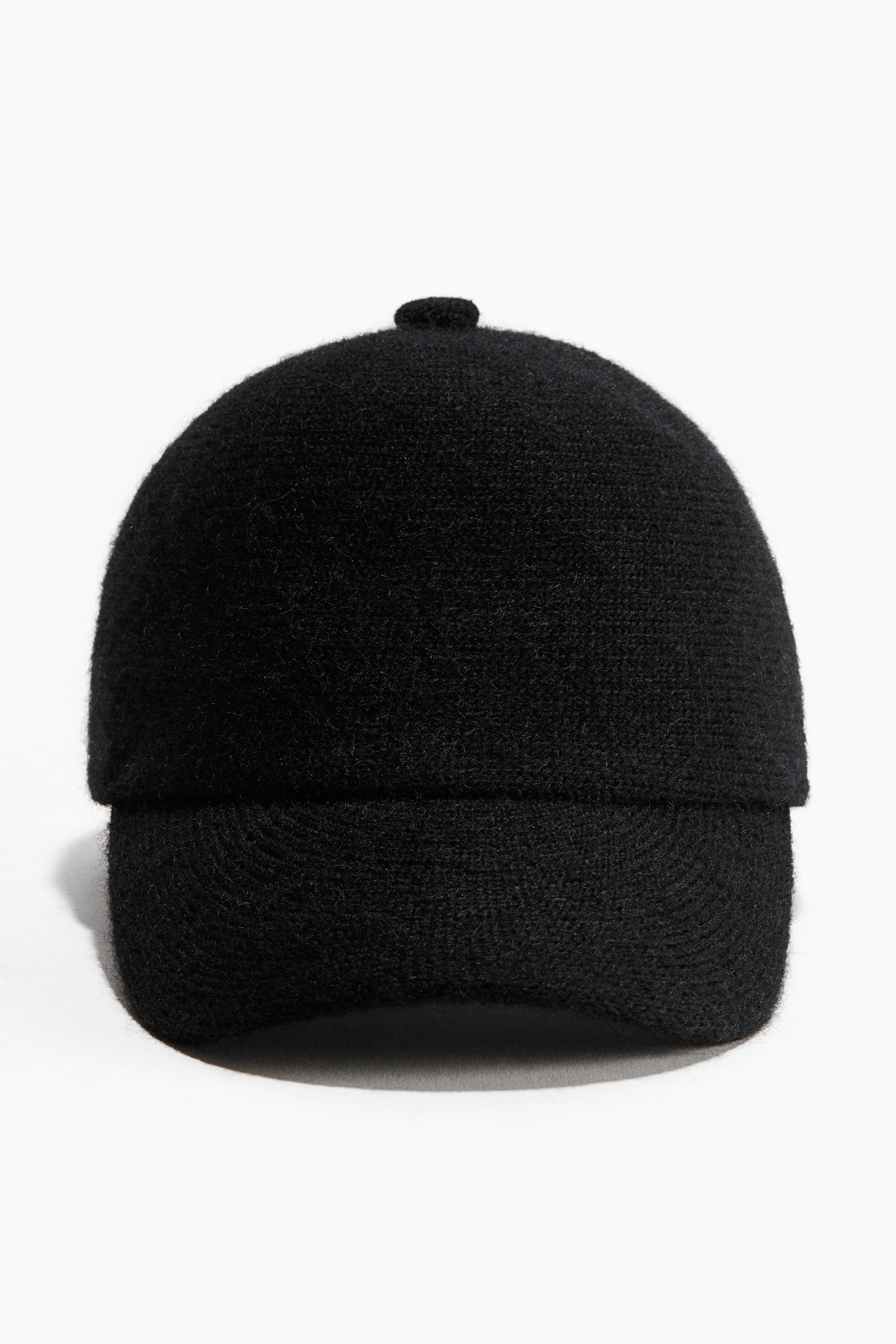 A black knit cap featuring a soft, textured finish and a curved brim is being worn by the model. The cap is styled alongside a matching black collared garment.