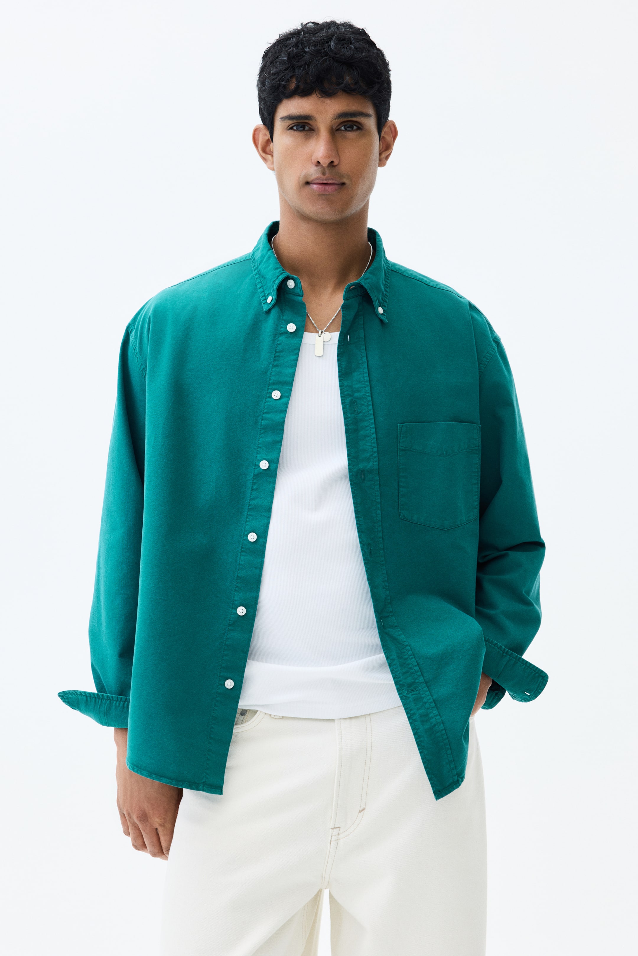 View larger image: The model is donning a teal Oxford cotton shirt, characterized by a button-down collar and an open chest pocket. It boasts a relaxed fit, with the long sleeves casually rolled up. The shirt is styled open over a white tank top, adding a touch of effortless elegance.