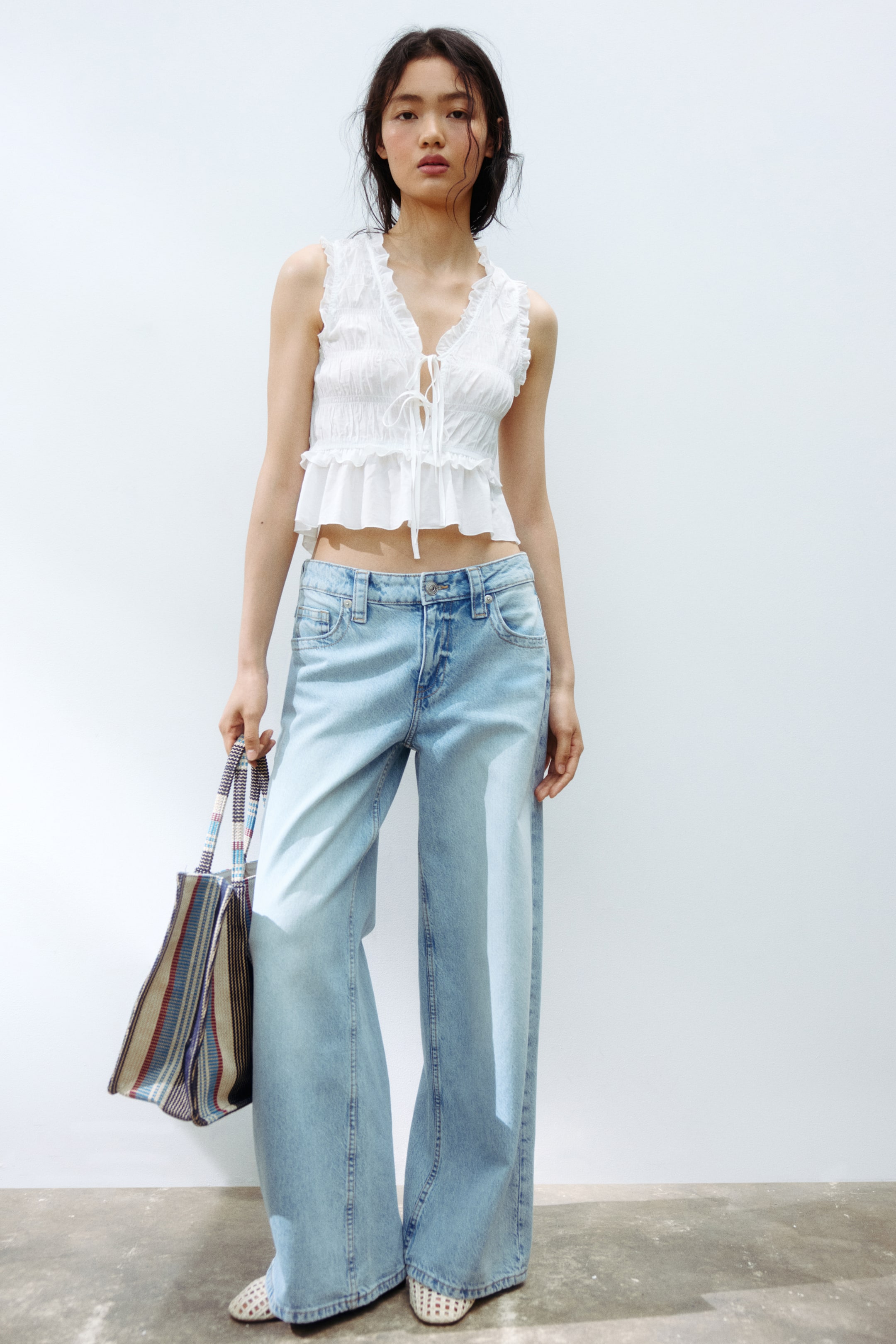 View larger image: Light wash 5-pocket jeans are worn by the model, showcasing a low waist with a visible button closure, and wide legs with a baggy fit from the thigh to the hem. The regular length hem rests on the top of the model's foot, and the model pairs the jeans with a white sleeveless top and carries a striped tote bag.