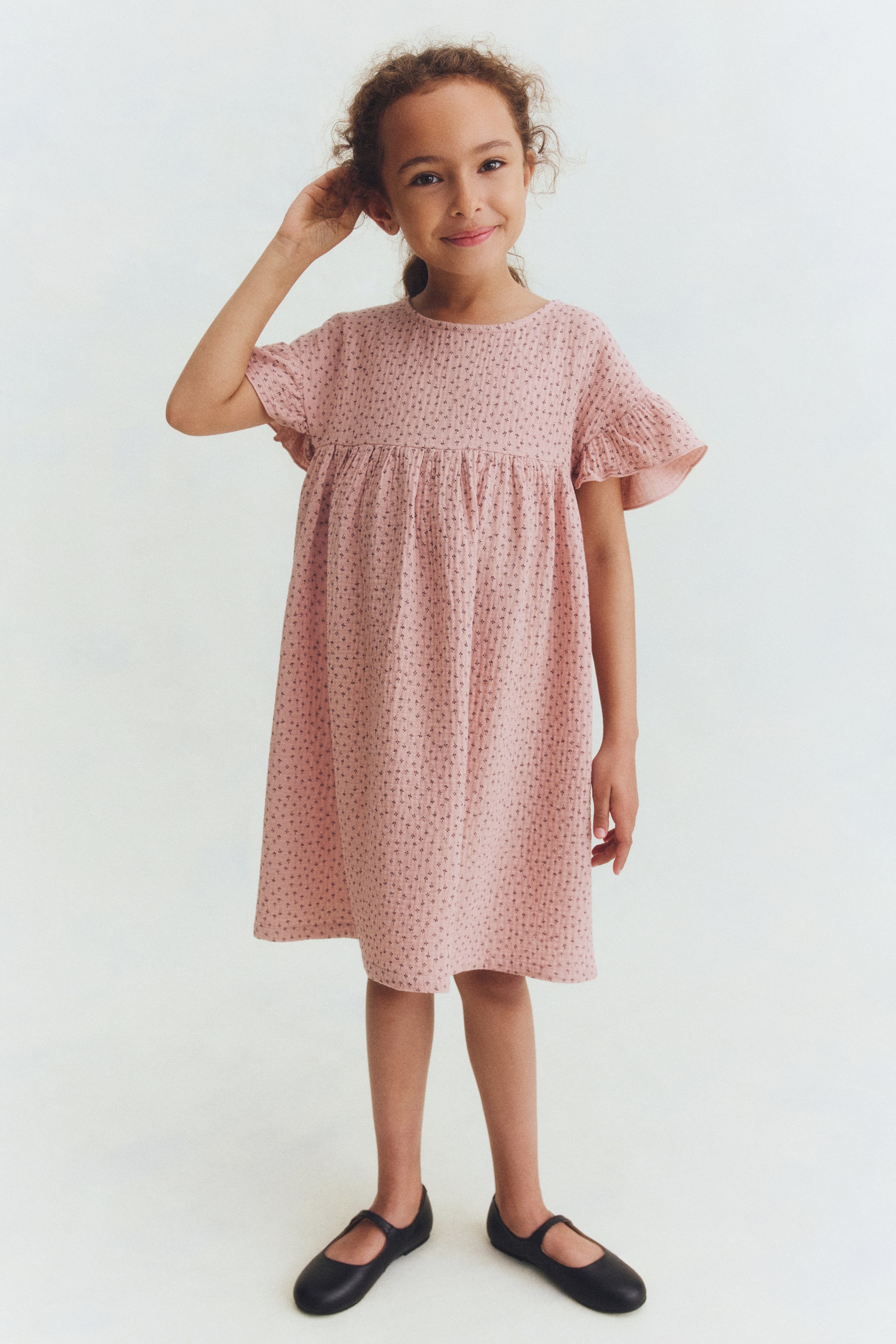 Ampliar la imagen: Un vestido rosa presenta una tela texturizada con un diminuto patrón oscuro en forma de cruz, un escote redondo y mangas cortas con volantes. El vestido se recoge en la cintura estilo imperio, fluyendo hacia una falda en línea A, y está posicionado ligeramente hacia la derecha.