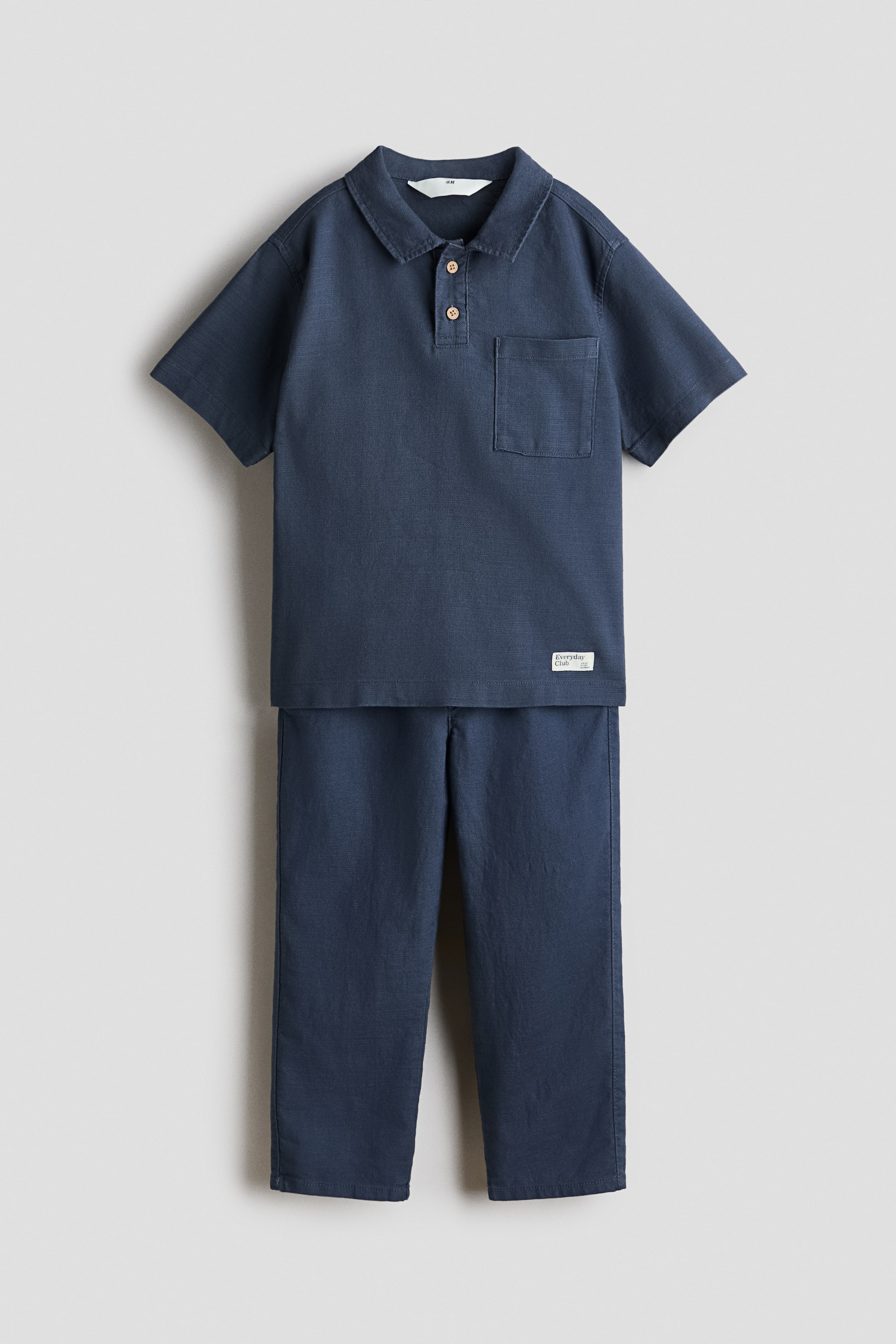 View larger image: Shown from the front, the navy blue two-piece set comprises a short-sleeved polo shirt featuring a collar, a two-button placket with light-brown buttons and a patch pocket on the left chest. Matching straight-leg trousers complete the outfit.