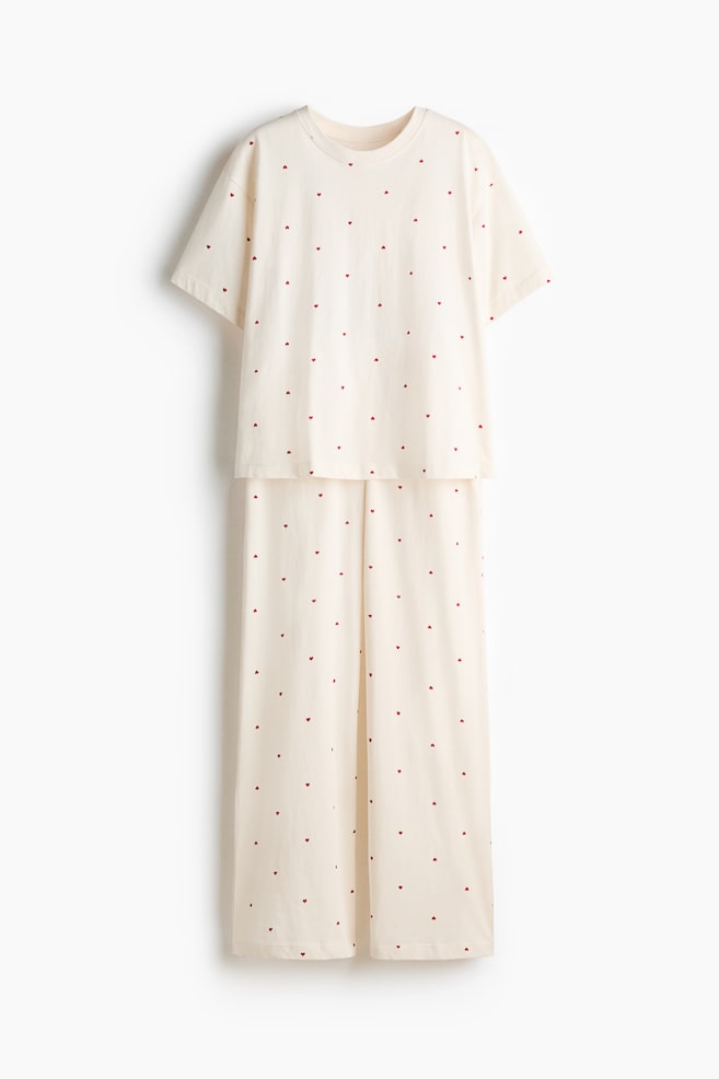A light cream pyjama set is presented front-on, featuring a T-shirt and full-length bottoms, both patterned with small red hearts. The T-shirt showcases a round, ribbed neckline, short sleeves and dropped shoulders, while the bottoms are designed with an elasticated waist and straight legs. - 1