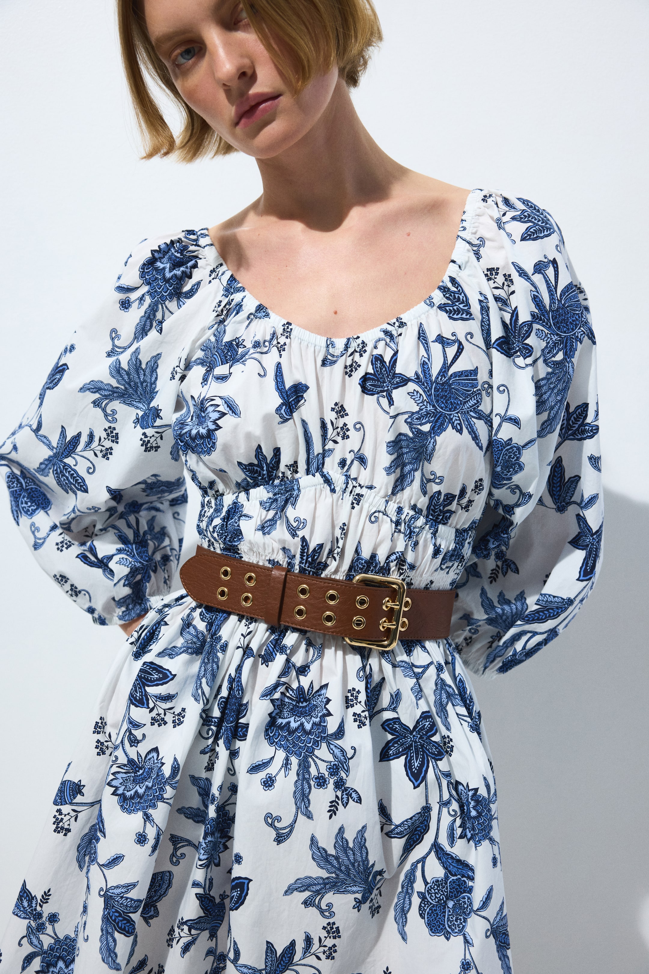 Ver imagem maior: Um vestido branco com um padrão floral e botânico azul apresenta um decote redondo franzido, mangas bufantes e uma cintura elástica. Um cinto castanho com ilhós dourados e uma fivela retangular está a apertar a cintura, criando uma saia fluida.
