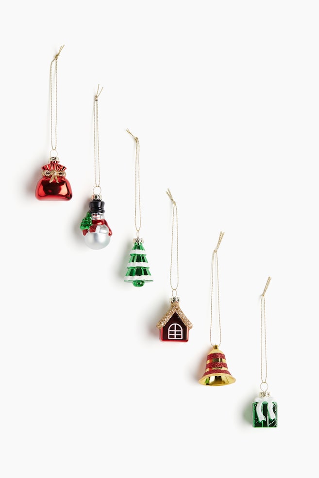 Plastikweihnachtsdekorationen zeigen verschiedene Designs, die jeweils eine Öse an der Oberseite mit einer glitzernden goldenen Kordel aufweisen. Die VORDERSEITE der Dekorationen zeigt eine glänzende rote Geschenktüte mit einer goldenen Schleife, einen Schneemann, der einen kleinen grünen Baum hält, einen grünen gestuften Weihnachtsbaum mit weißen Details, ein glänzendes rotes Haus mit einem glitzernden braunen Dach und einem weißen Fensterrahmen, eine goldene Glocke mit roten horizontalen Streifen und eine glänzende grüne Geschenkbox mit einem weißen Band und Schleife. - 1