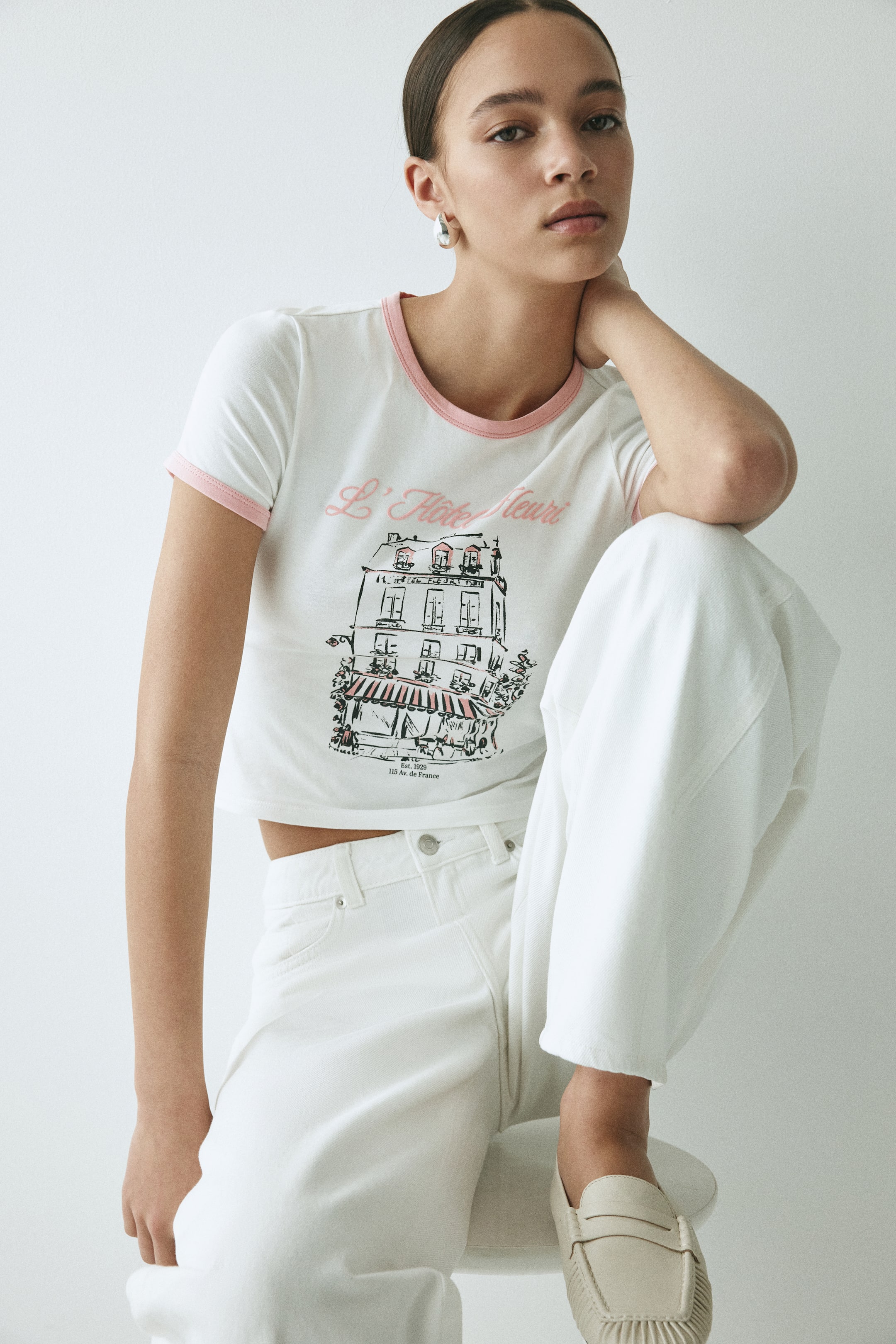 View larger image: A white cropped T-shirt features a round neckline and short sleeves, both finished with pink ribbed trim. The front displays a graphic print reading "L'Hôtel Fleuri" in pink script, centred above a line drawing of a multi-storey building with pink awnings. Smaller text reading "Est. 1929" and "115 Av. de France" is visible below the building.