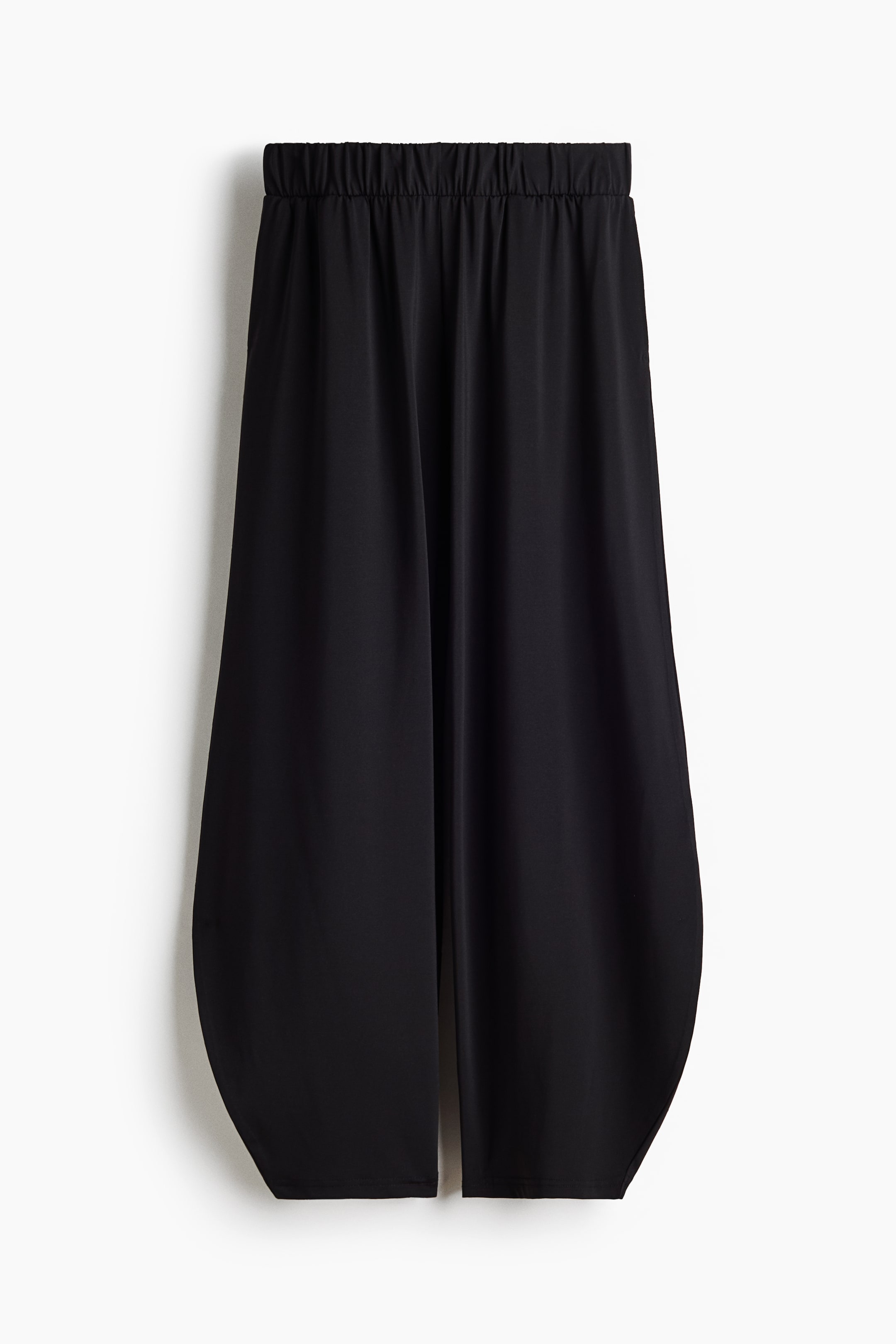 View larger image: Black trousers are showcased in a front view, featuring a gathered elasticated waistband. The wide, flowing legs culminate in a distinct curved hemline that ascends towards the center of each leg.
