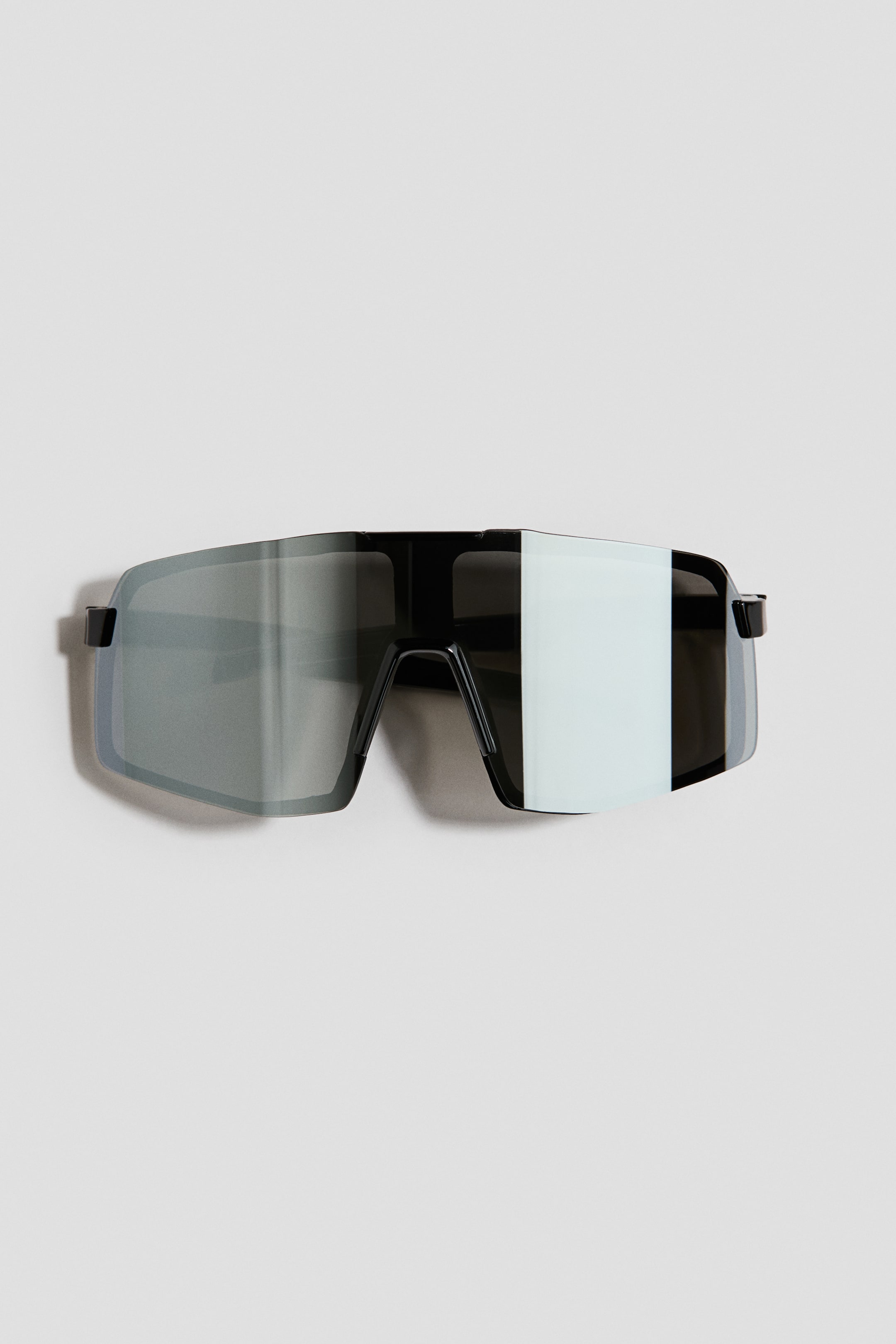 Grösseres Bild ansehen: Die Sonnenbrille ist frontal ausgerichtet und hat übergrosse, getönte sowie verspiegelte Visiergläser. Ein schwarzer Rahmen geht oben und an den Seiten in die Gläser über.