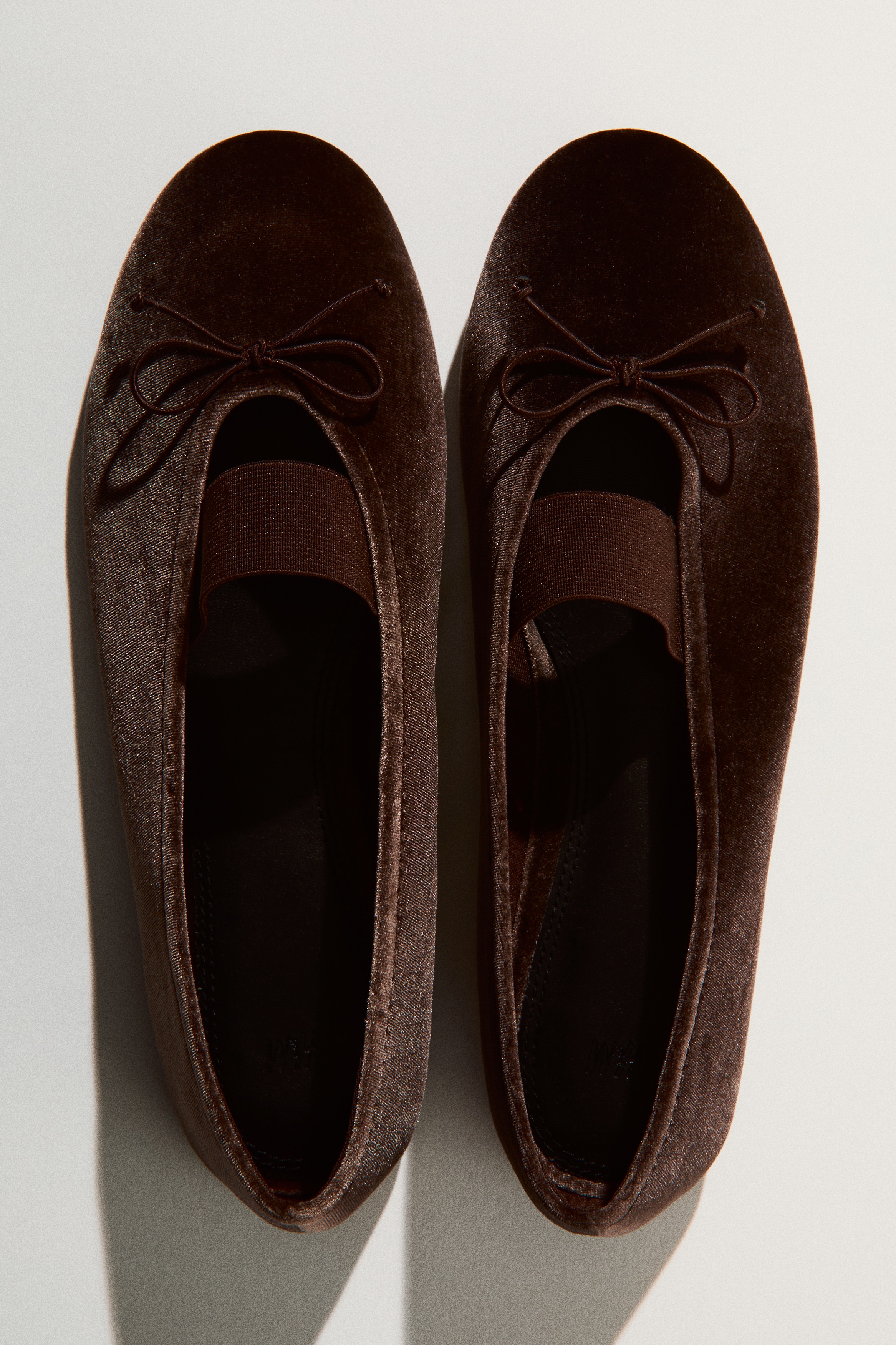 A pair of dark brown velvet ballet flats is being showcased, highlighting the front toe and instep. Each flat is adorned with a small bow tie on the toe and an elastic strap stretching across the instep. The black insole displays the H&M logo.