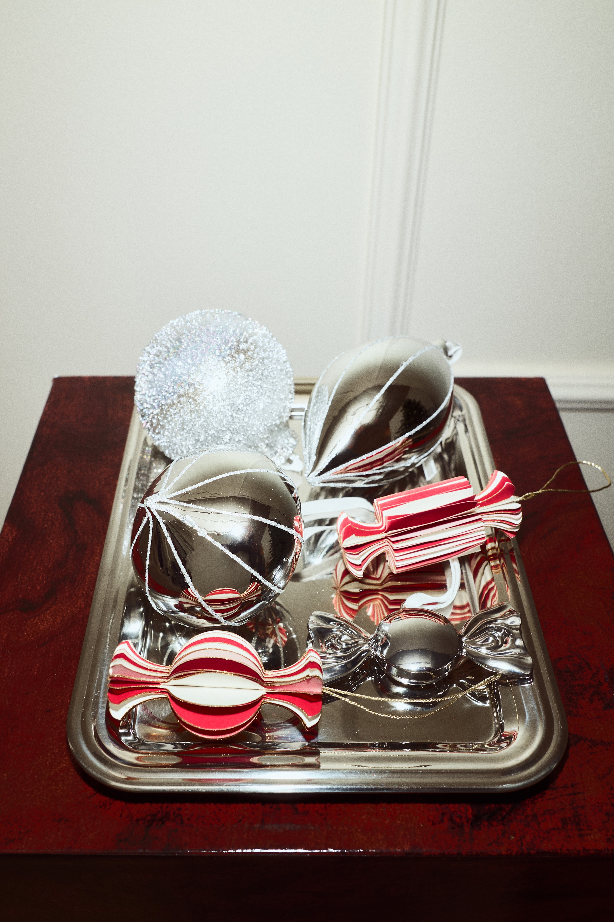 Two front-facing paper ornaments shaped like wrapped candies hang from thin gold strings, displaying a three-dimensional, accordion-folded structure with alternating red and cream stripes and gold-glittered edges. The left ornament showcases a spherical centre, while the right ornament features a cylindrical centre, with both designs finishing in flared ends.