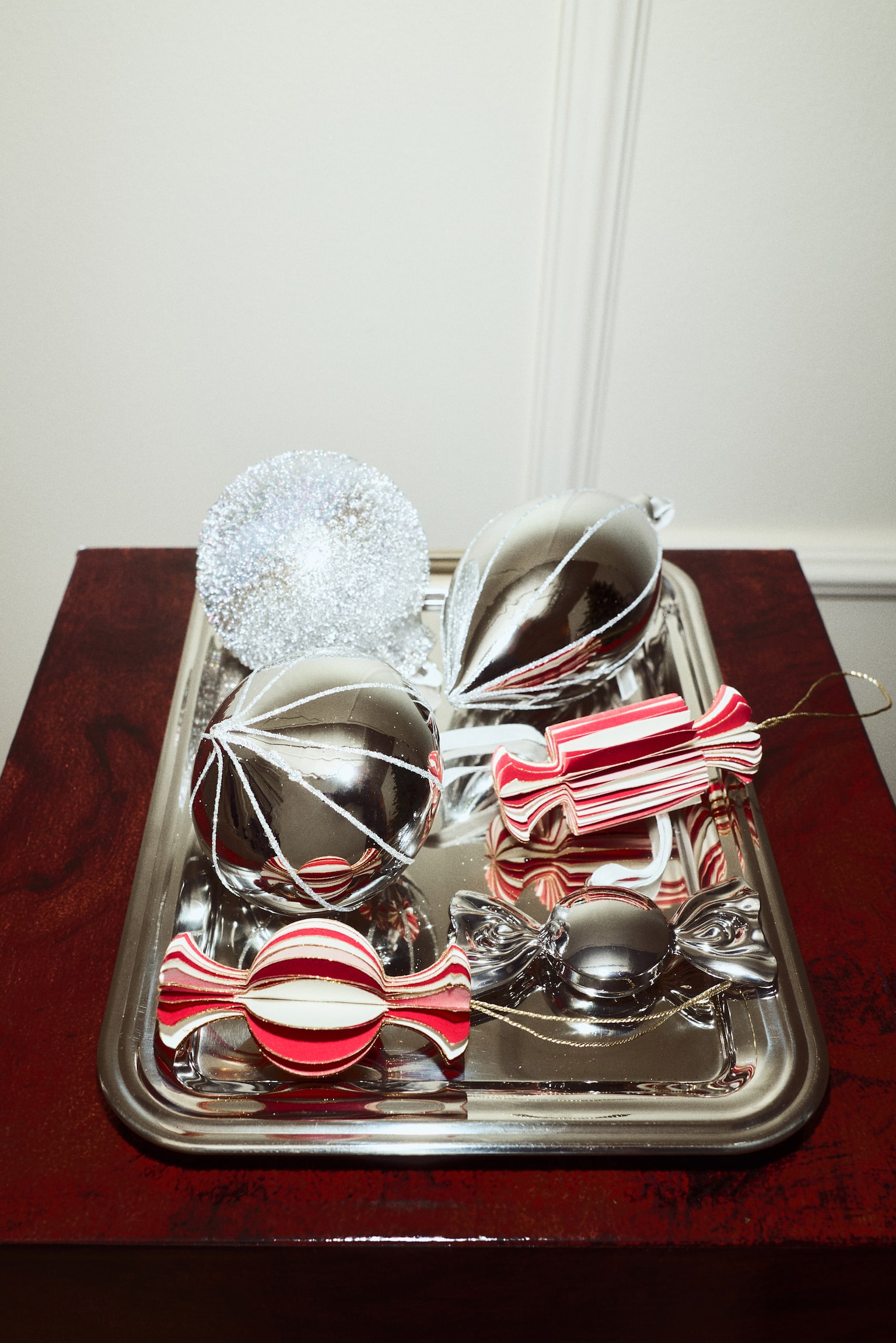 Two front-facing paper ornaments shaped like wrapped candies hang from thin gold strings, displaying a three-dimensional, accordion-folded structure with alternating red and cream stripes and gold-glittered edges. The left ornament showcases a spherical centre, while the right ornament features a cylindrical centre, with both designs finishing in flared ends. - 1