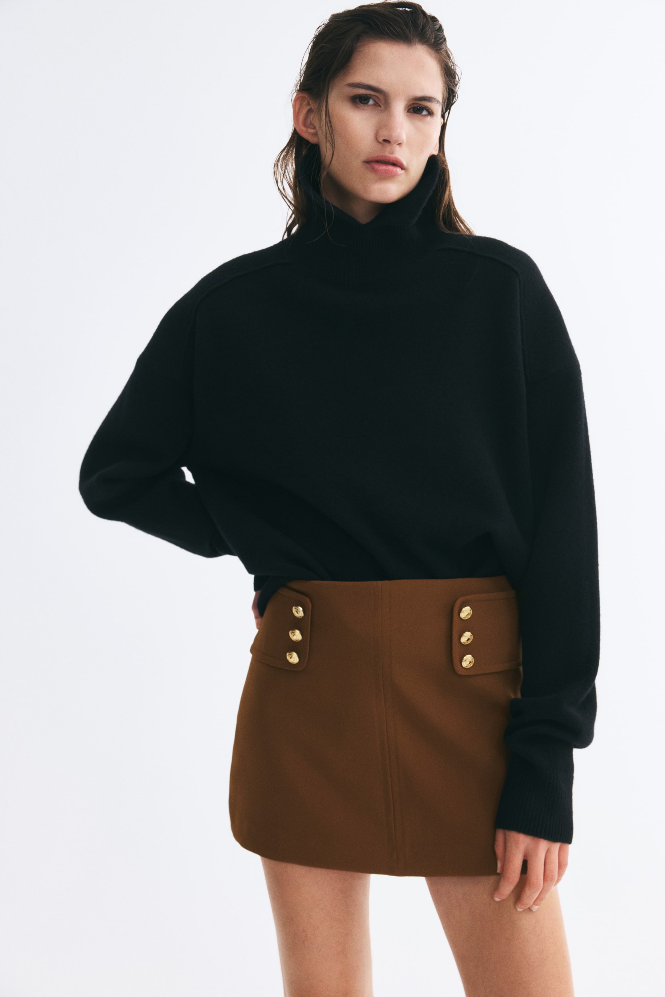 Ampliar la imagen: La modelo lleva una falda-pantalón corta color café, con amplias lengüetas laterales a la cintura detalladas con botones decorativos color dorado y una costura visible en el centro del frente. La falda-pantalón se combina con un suéter negro de cuello alto y mangas largas, metido por dentro de la pretina.