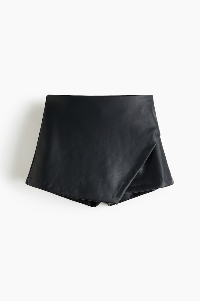 Las faldas pantalón de piel sintética negra están extendidas, mostrando una textura suave y una pretina ancha y plana. Un panel frontal superpuesto crea una apariencia similar a una falda. - 1