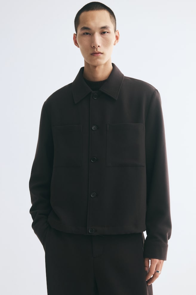 The model is wearing a dark brown overshirt in a soft weave over a black crew-neck top and matching trousers. The overshirt features a pointed collar, front button fastening, dropped shoulders, and two large patch chest pockets. - 1