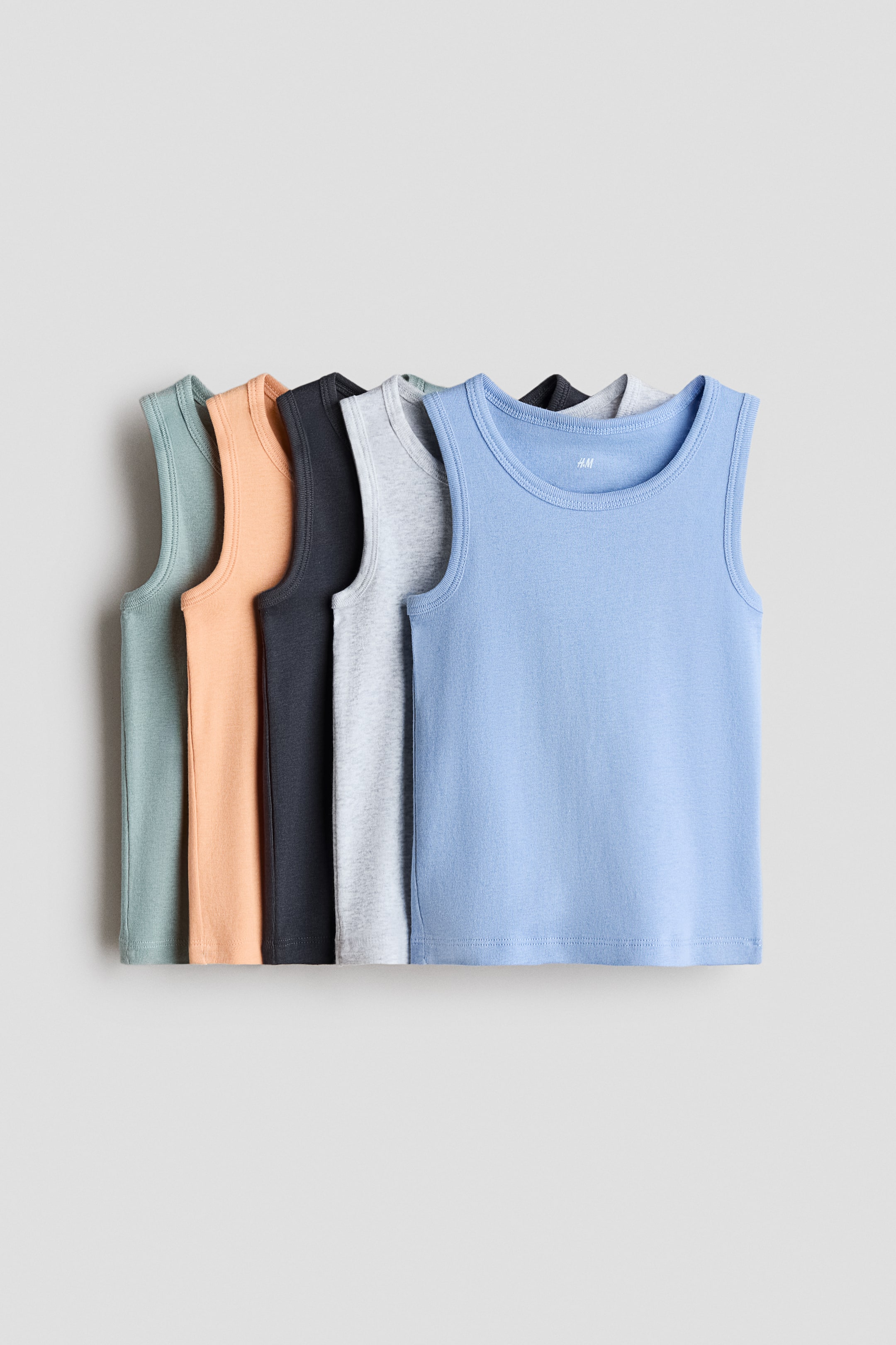 View larger image: Vest tops are neatly lined up, showcasing their fronts. They are crafted from soft cotton jersey material, complete with ribbed trim at the neckline and armholes. Available in solid hues such as light blue, light grey, dark grey, peach, and sage green.