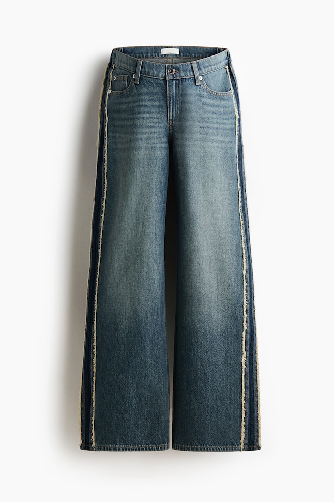 The wide-leg jeans are facing forward, showcasing a medium blue wash with lighter fading on the thighs. They feature a button and zip fastening at the waist, front pockets, and a darker denim stripe bordered by frayed edges running down each outer leg. - 1