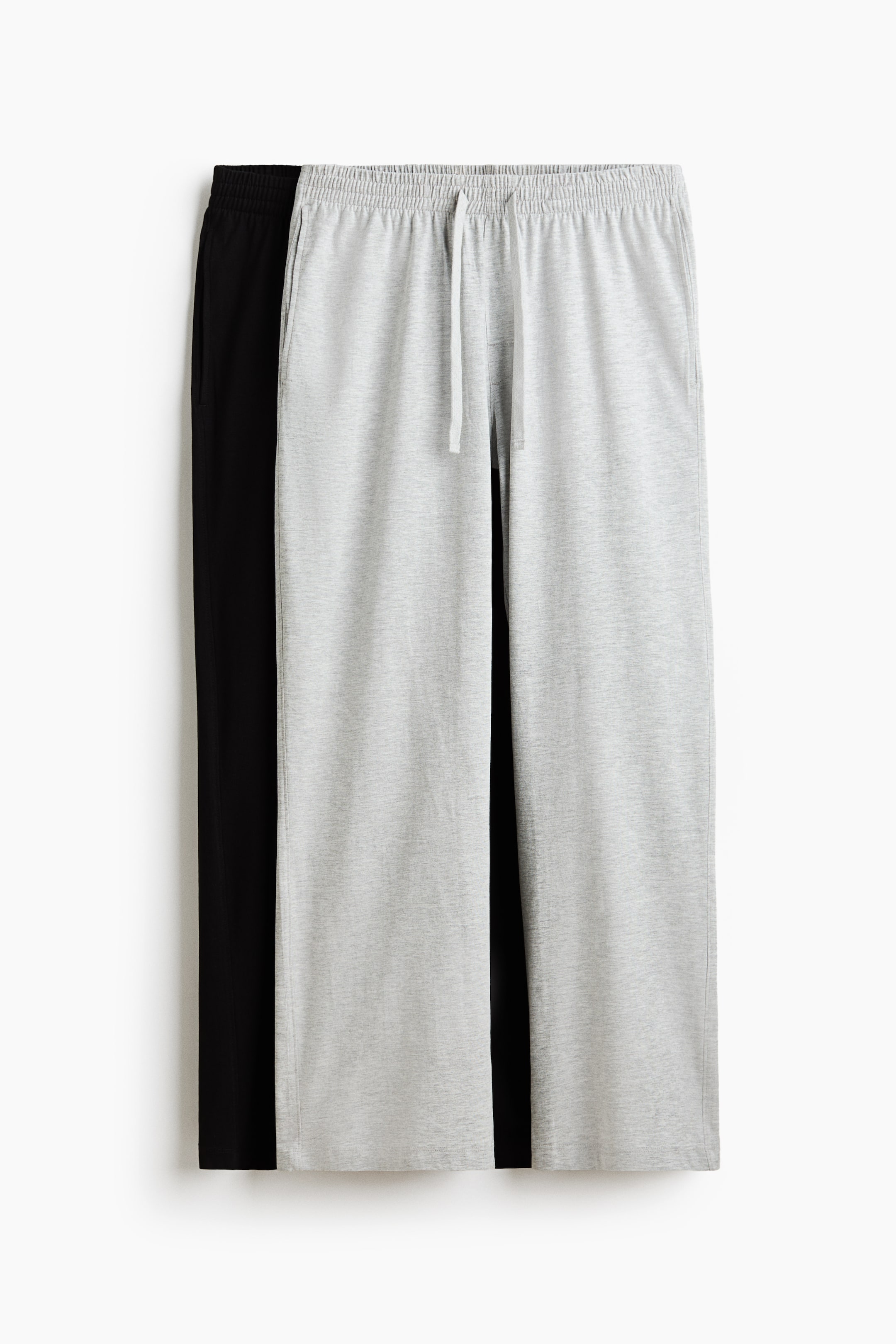 Größeres Bild ansehen: Zwei Paar Jogginghosen, eine hellgraue und eine schwarze, werden präsentiert. Die hellgraue Hose, die nach vorne zeigt, verfügt über einen elastischen Bund mit weißem Kordelzug und Seitentaschen. Die schwarze Hose ist teilweise dahinter zu sehen.