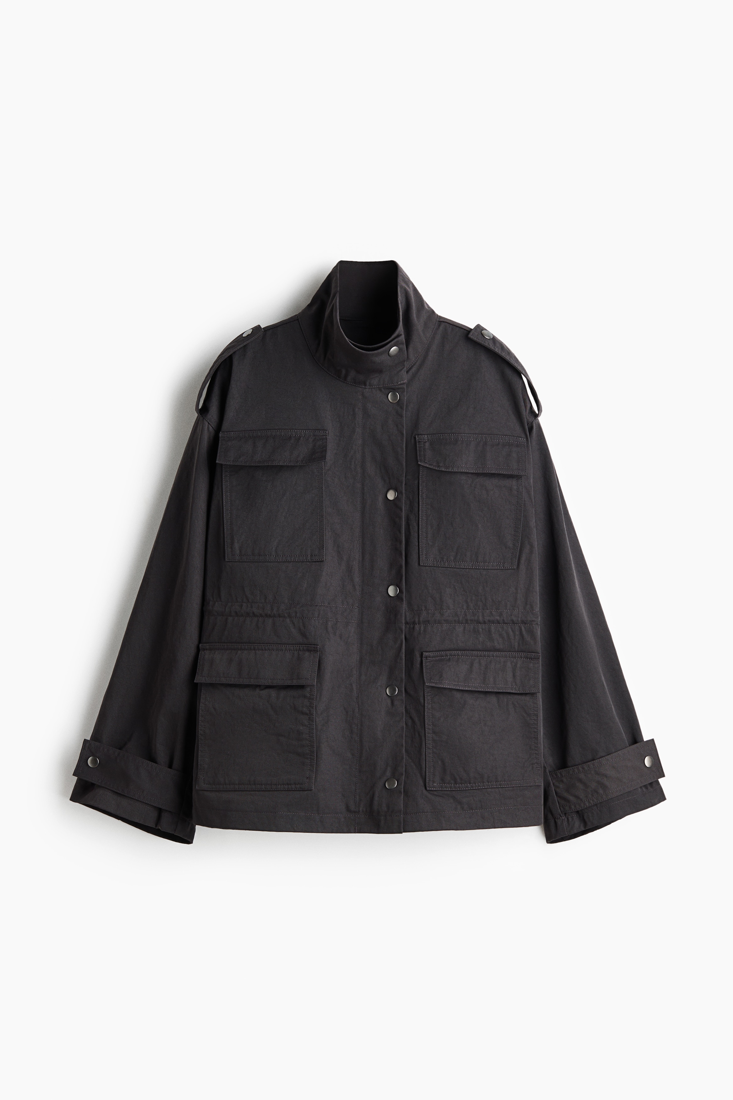 An oversized black jacket is being worn by the model, featuring a high funnel collar, press-stud closures down the front, shoulder tabs, and large flap pockets on the chest and hips. The jacket is cinched at the waist with a drawstring and layered over a cream-colored skirt.