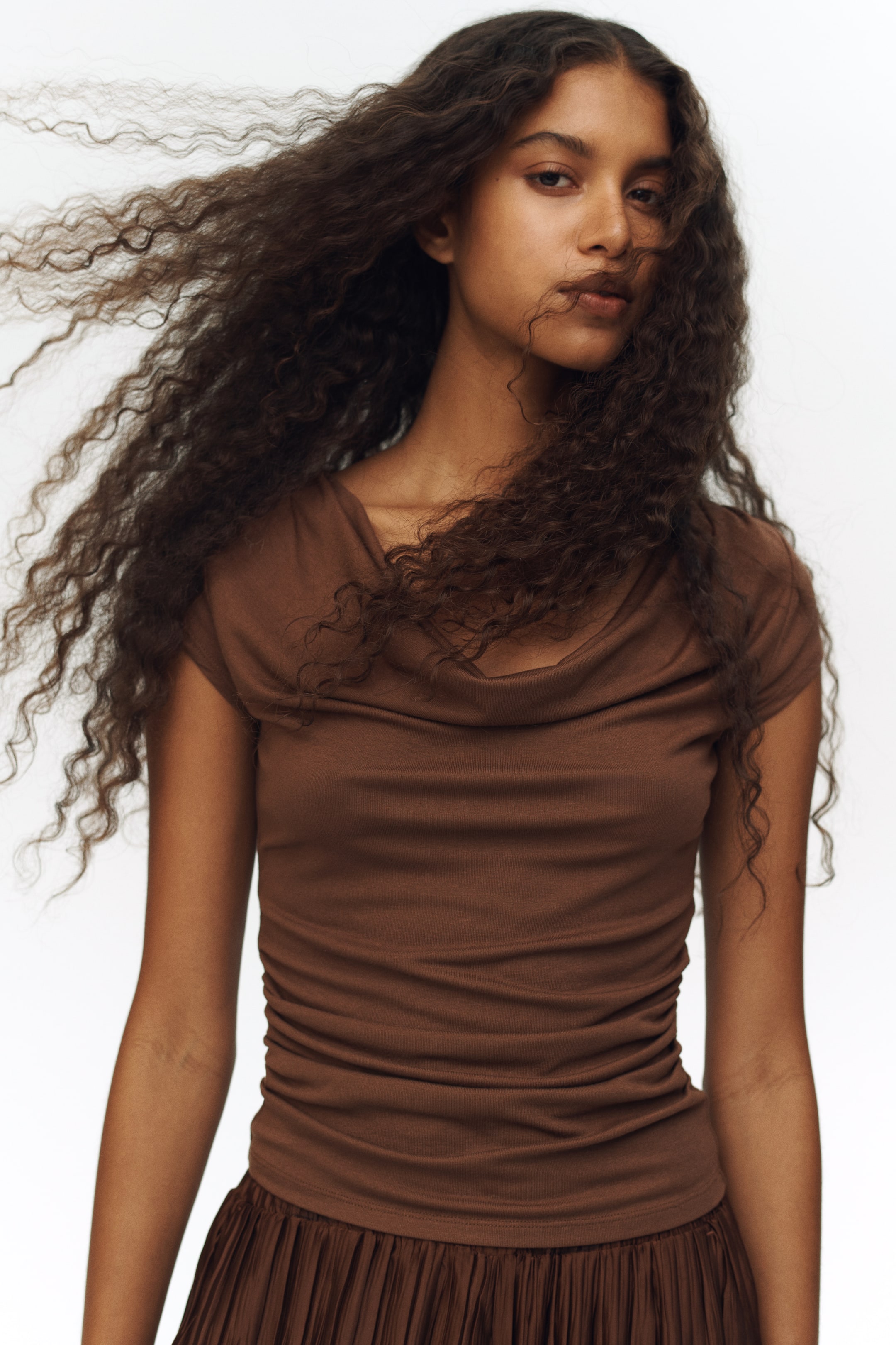 View larger image: The brown top is displayed front-facing, highlighting a draped cowl neck and short sleeves. It fits snugly to the torso with side ruching and is paired with a pleated brown skirt.