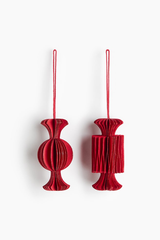 Two red paper ornaments shaped like wrapped candies are hanging side by side, facing forward. The left ornament displays a rounded, spherical centre with flared ends, while the right ornament displays a rectangular, pleated centre, with both featuring layered accordion-style folds and hanging from thin red string loops. - 1