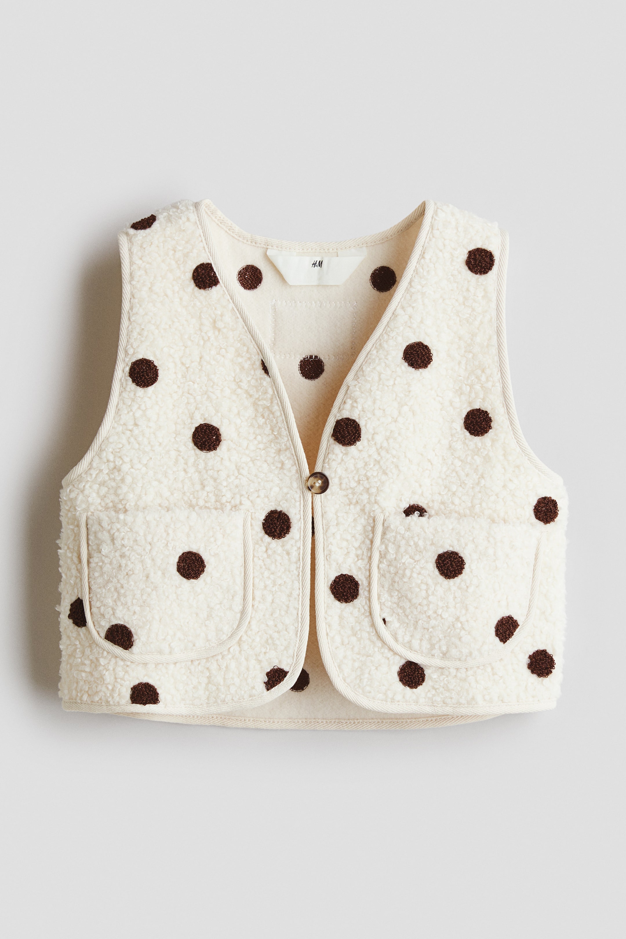 View larger image: A cream-colored sherpa vest is shown from the front, featuring an all-over brown polka dot pattern. It has a V-neckline and two patch pockets on the front, with a single brown button securing the opening, and cream-colored fabric binding accentuating all edges.
