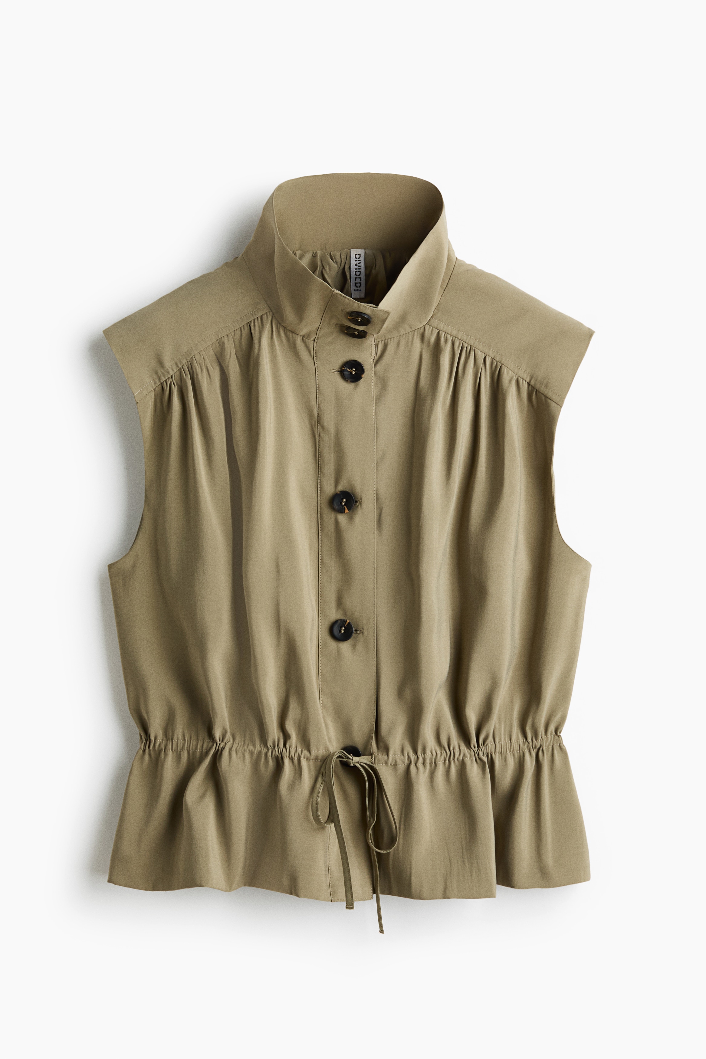 Het model draagt een mouwloze blouse in saliegroen, in trenchcoatform met een hoge opstaande kraag met knopen, een knoopsluiting vooraan en een gerimpelde taille met trekkoord. De blouse is gestyled met bijpassende wijde broekspijpen in dezelfde lichte stof.