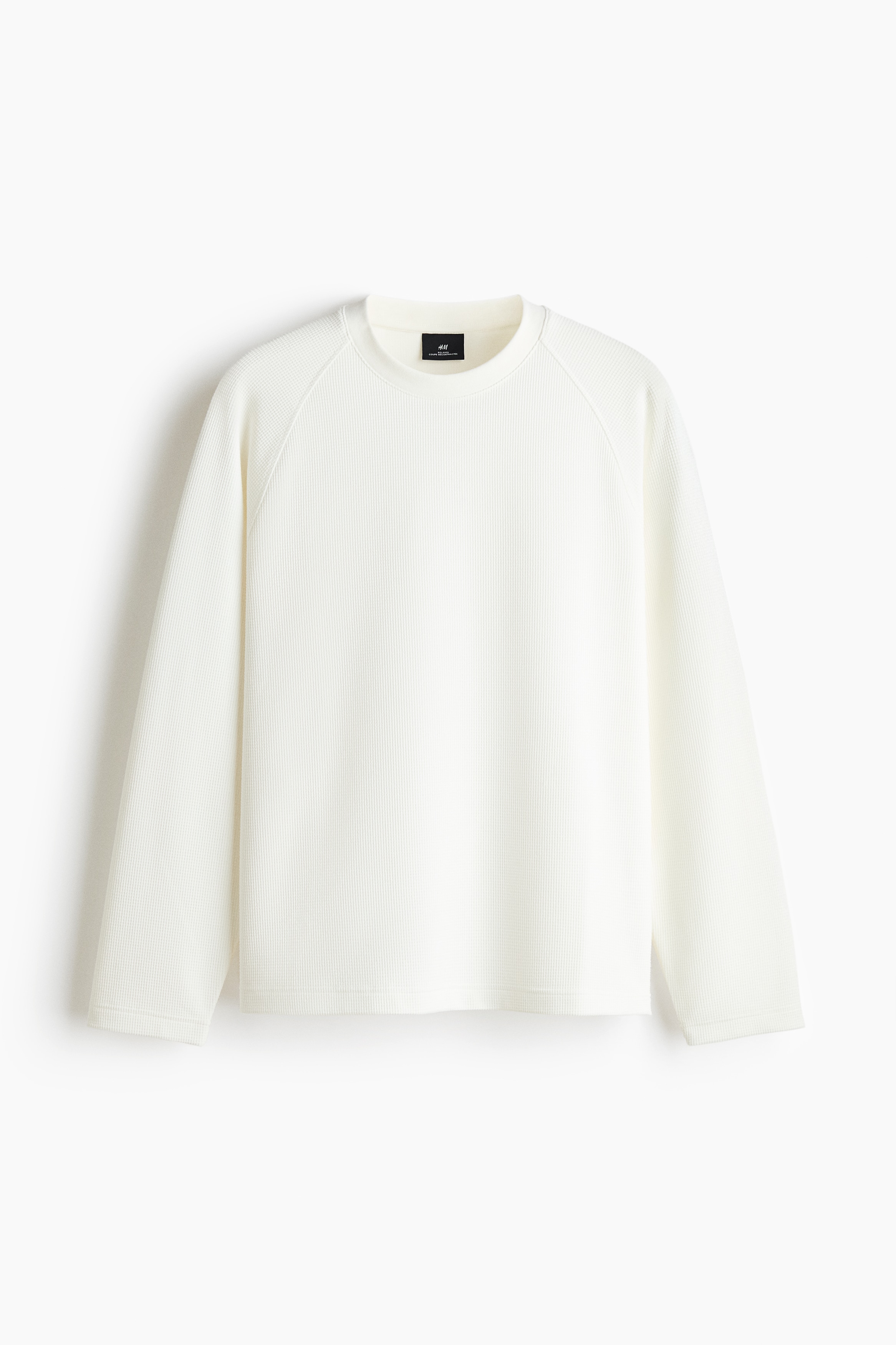 Un tee-shirt en jersey gaufrÃ© blanc cassÃ©, dotÃ© dâun col rond, de manches raglan longues et dâun ourlet droit, est portÃ© par le modÃ¨le. Le modÃ¨le se tient debout, une main glissÃ©e dans la poche dâun pantalon bleu foncÃ©, mettant en valeur la coupe dÃ©contractÃ©e et la silhouette casual de la chemise.