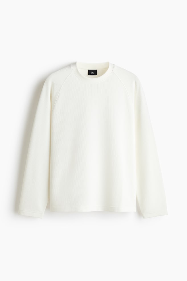 A cream-colored, waffle-knit long-sleeve shirt is shown facing front, featuring a ribbed crew neckline and long raglan sleeves. The garment is displaying a textured pattern throughout, with a straight hem and clean-finished cuffs. - 1