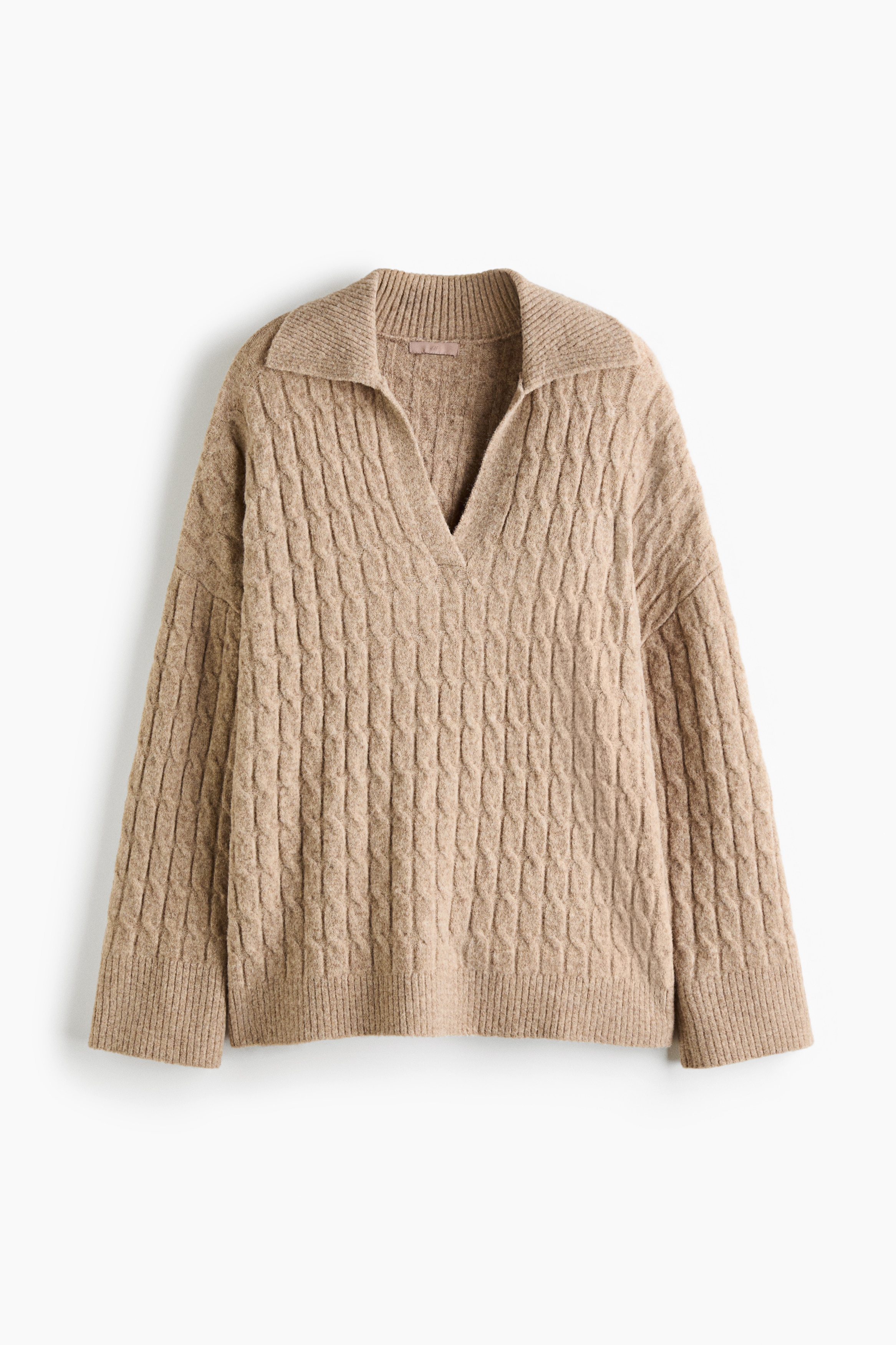 Ein oversized beiger Zopfpullover wird vom Model getragen und verfÃ¼gt Ã¼ber einen gerippten offenen Kragen, Ã¼berschnittene Schultern sowie einen gerippten Saum mit Seitenschlitzen. Der Pullover wird mit weiÃen Shorts gestylt, wÃ¤hrend das Model die HÃ¤nde in die HÃ¼ften stÃ¼tzt.