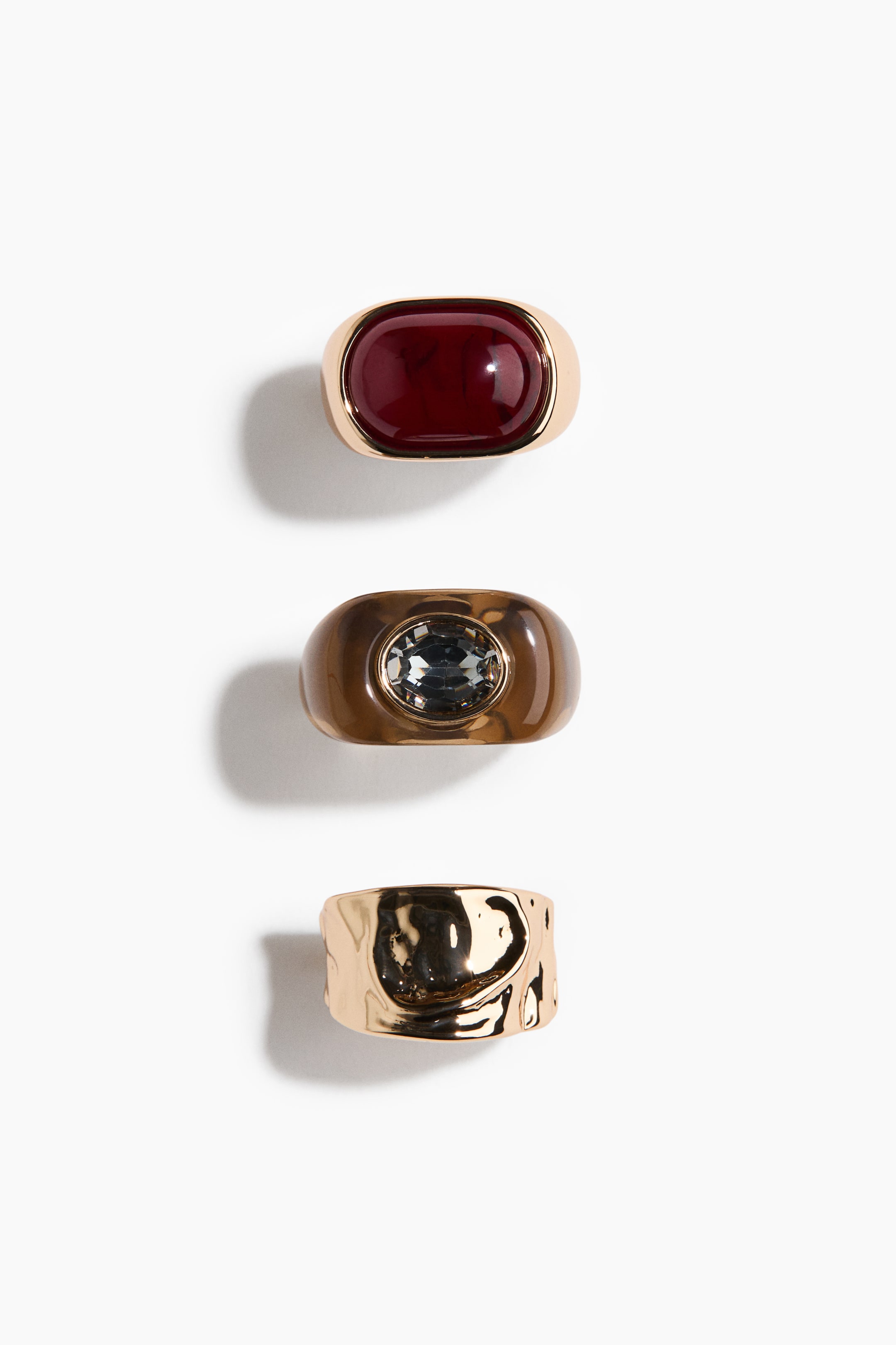 Größeres Bild ansehen: Drei Ringe liegen flach und präsentieren ihre Oberflächen. Der erste Ring ist ein goldfarbener Band mit einem ovalen, dunkelroten Cabochon-Stein, der zweite ist ein breites, braunes Band mit einem ovalen, facettierten grauen Stein, und der dritte ist ein breites, goldfarbenes Band mit einer unregelmäßigen, strukturierten Oberfläche.