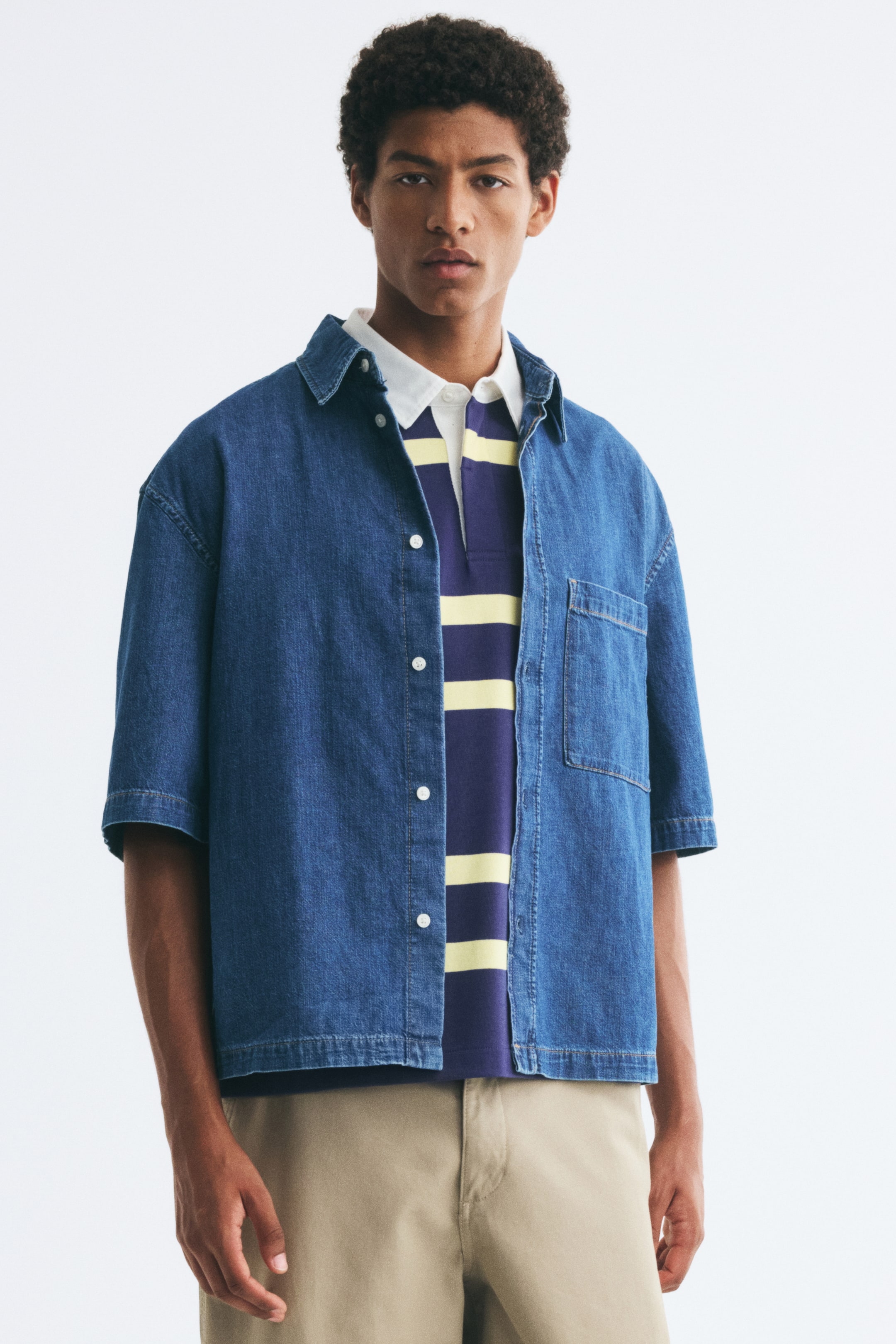 Xem ảnh lớn hơn: Người mẫu đang mặc áo sơ mi denim tay ngắn mở cúc, với phom dáng thoải mái, cổ áo cổ điển, vai trễ và một túi ngực đắp đơn. Áo được phối lớp bên ngoài áo polo kẻ sọc và kết hợp cùng quần dài màu be.
