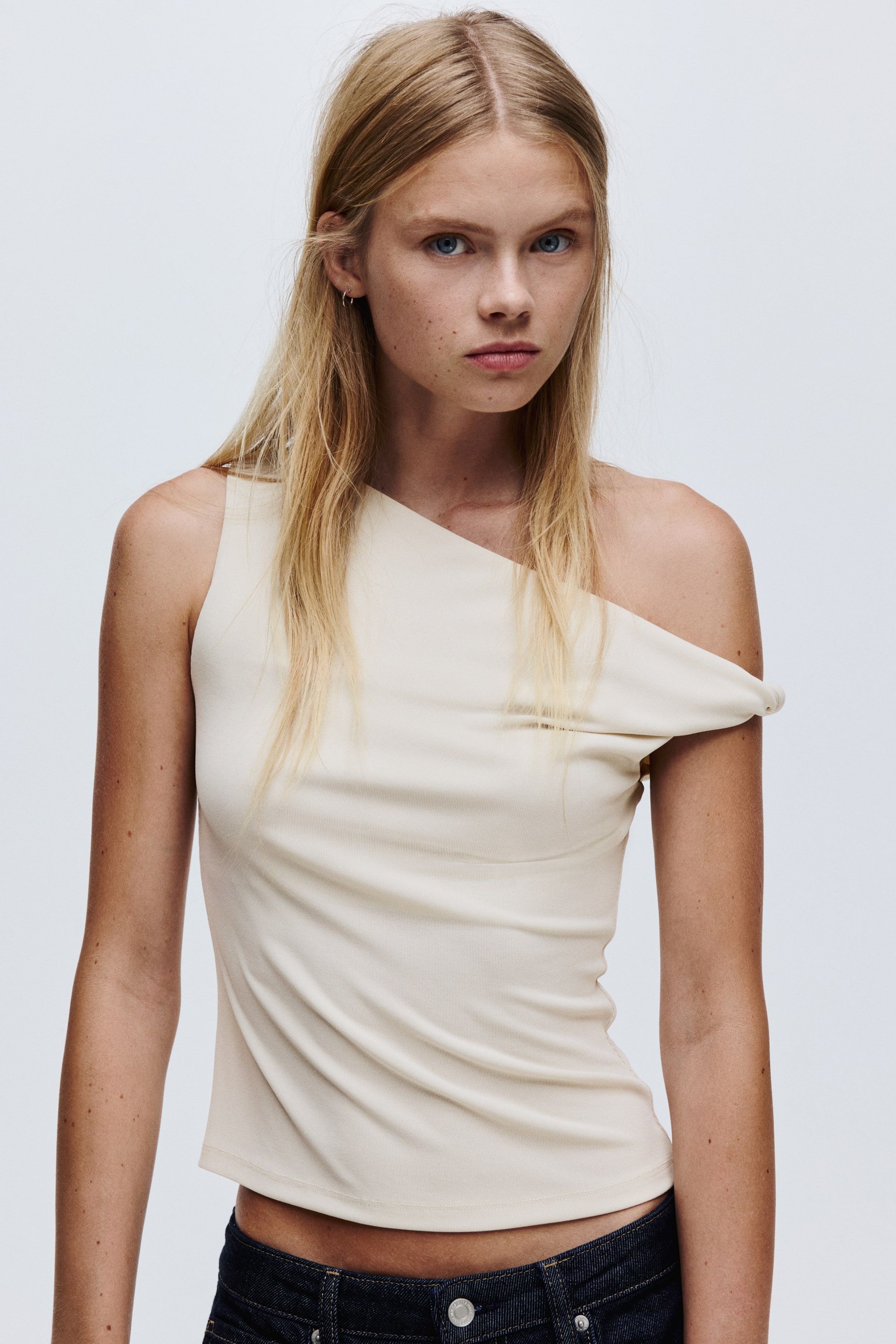 View larger image: A cream one-shoulder top is being presented front-facing, featuring an asymmetrical neckline with a strap on the left and a draped off-the-shoulder design on the right. The top is fitting closely to the body and ending above the waist.
