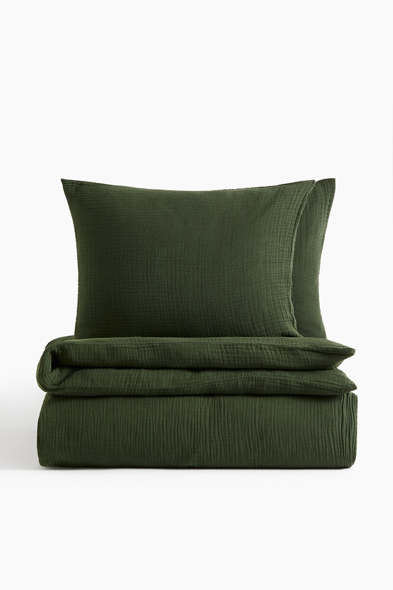 Ett mÃ¶rkgrÃ¶nt bÃ¤ddset i bomullsmuslin visas framifrÃ¥n, med ett prydligt vikt pÃ¥slakan och tvÃ¥ matchande kuddfodral som stÃ¥r upprÃ¤tt bakom det. Setet visar en skrynklig, texturerad muslinfinish.