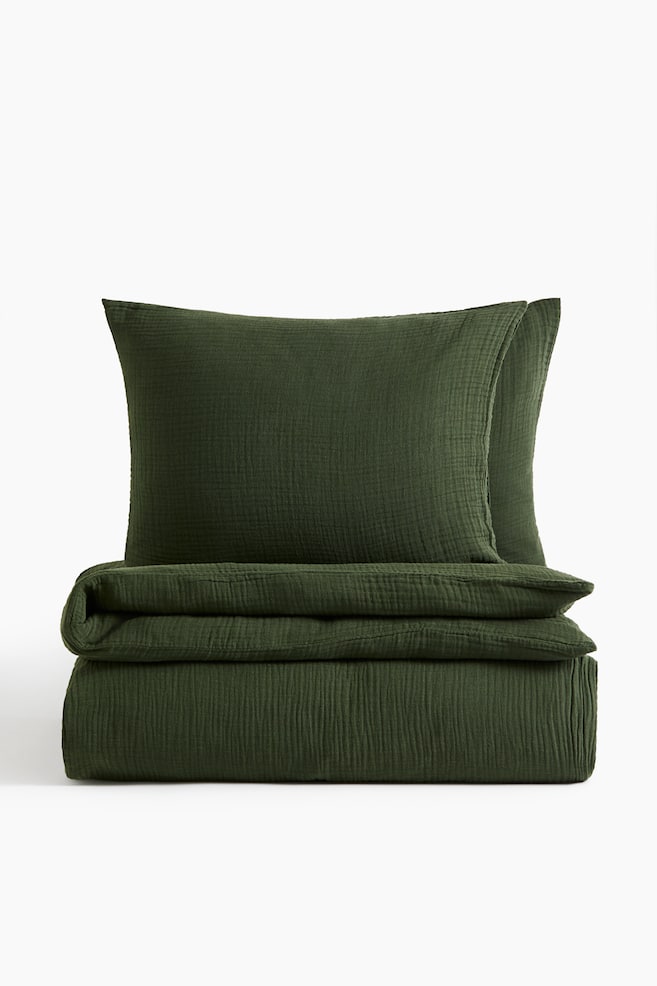 Un ensemble de housse de couette en mousseline de coton vert foncé est présenté de face, avec une housse de couette soigneusement pliée et deux taies d'oreiller assorties dressées derrière. Lâensemble met en valeur une finition en mousseline froissée et texturée. - 1