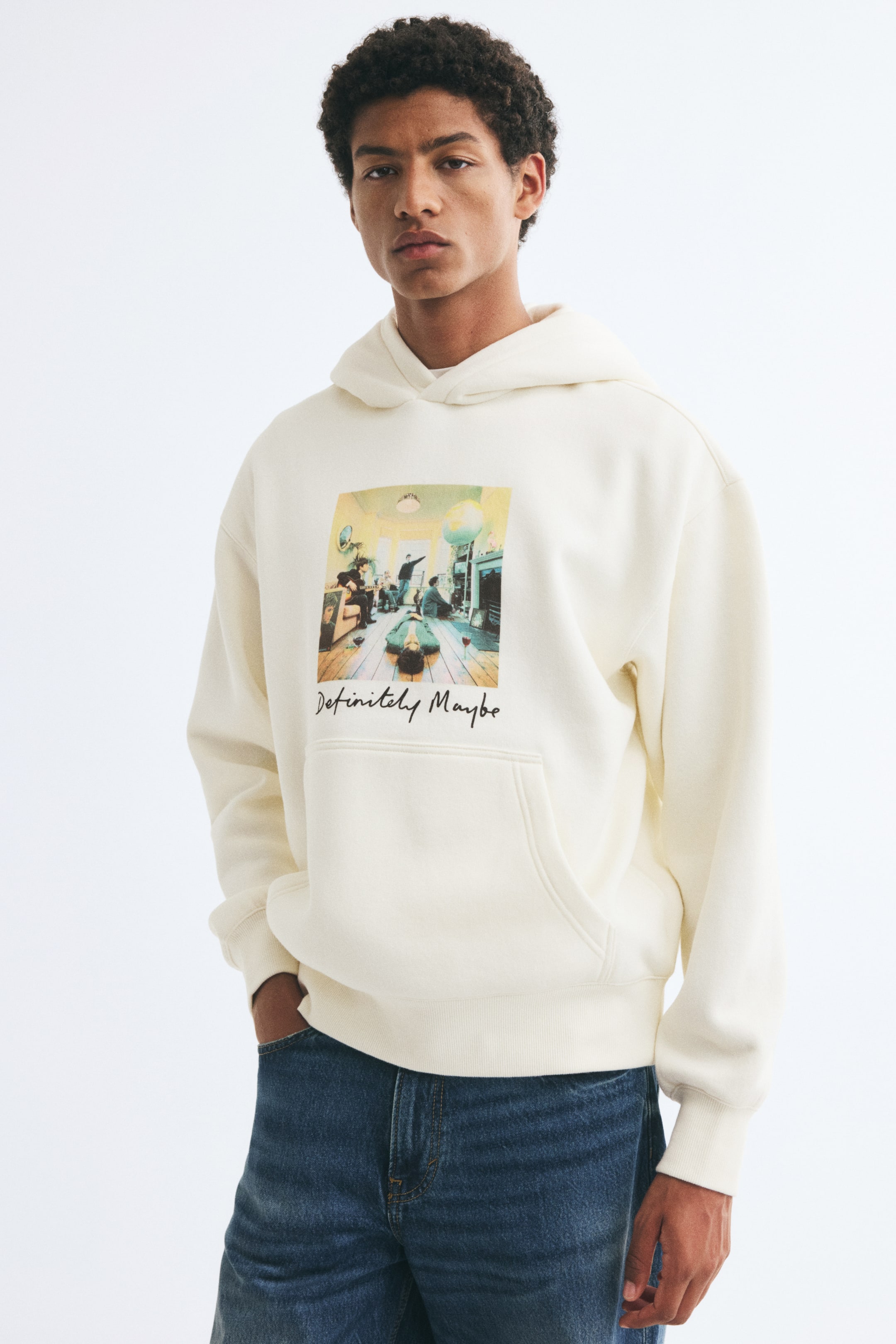 View larger image: The model is wearing a cream-colored hoodie featuring a graphic print of the Oasis "Definitely Maybe" album cover on the chest. The hoodie is designed with a wrap hood, dropped shoulders, a kangaroo pocket, and ribbed trims, and is styled with blue jeans.