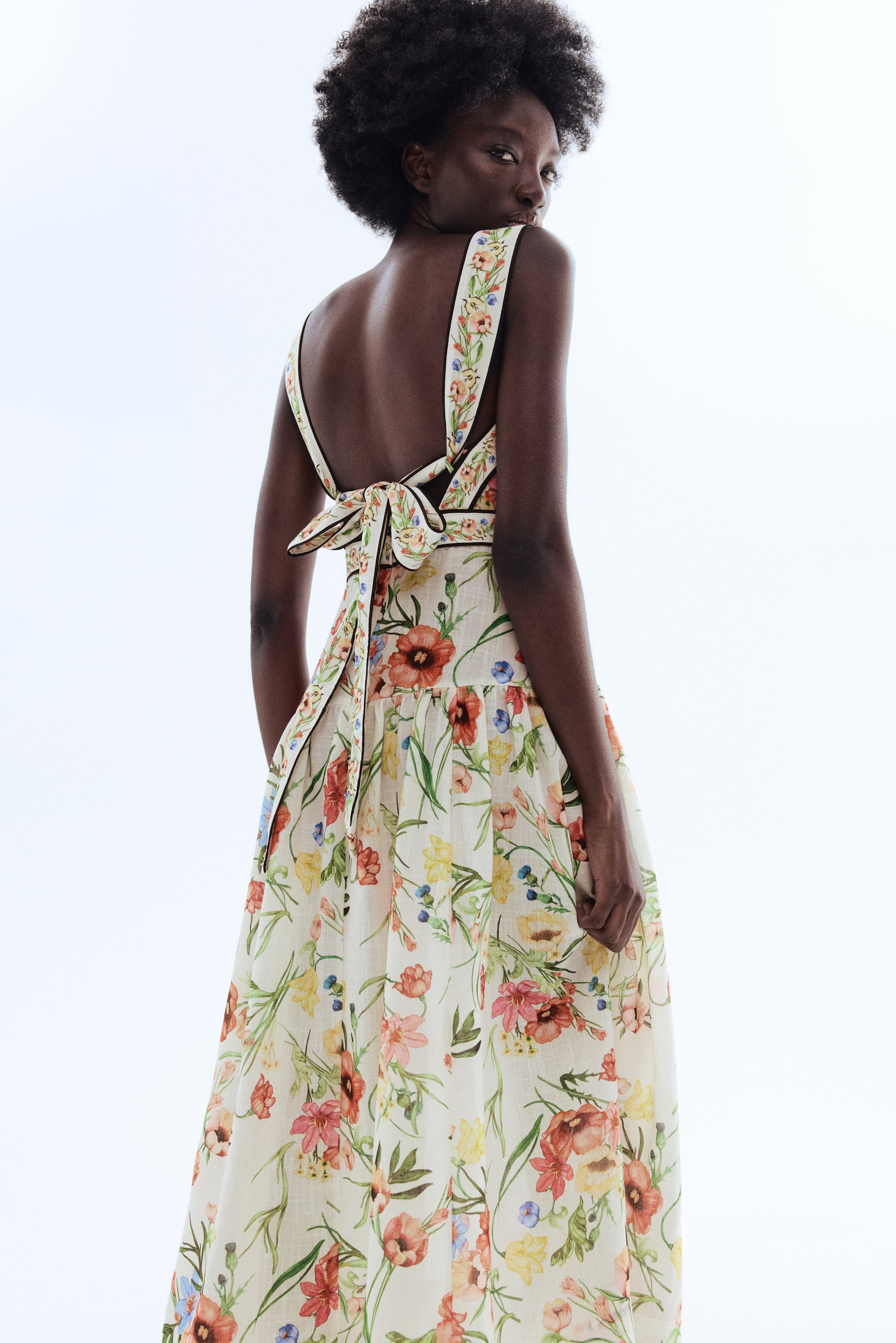 拡大画像を表示: 背面から斜め左に向けて撮影されたフローラルマキシドレスです。広めの花柄ストラップはダークなトリムが施され、オープンな上部の背中で大きなリボンに結ばれています。スカートは、明るい背景に鮮やかな花柄が広がるフルフローイングデザインです。