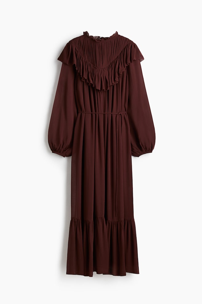 Ein braunes langärmliges Kleid wird von vorne präsentiert, mit einem gerafften hohen Kragen, einem V-förmigen, mit Biesen versehenen Mieder und einem großen Rüschenüberzug, der über die Schultern reicht. Das Kleid ist in der Taille mit einem Band gebunden und wird durch Ballonärmel und einen gestuften Rüschen-Saum vervollständigt. - 1