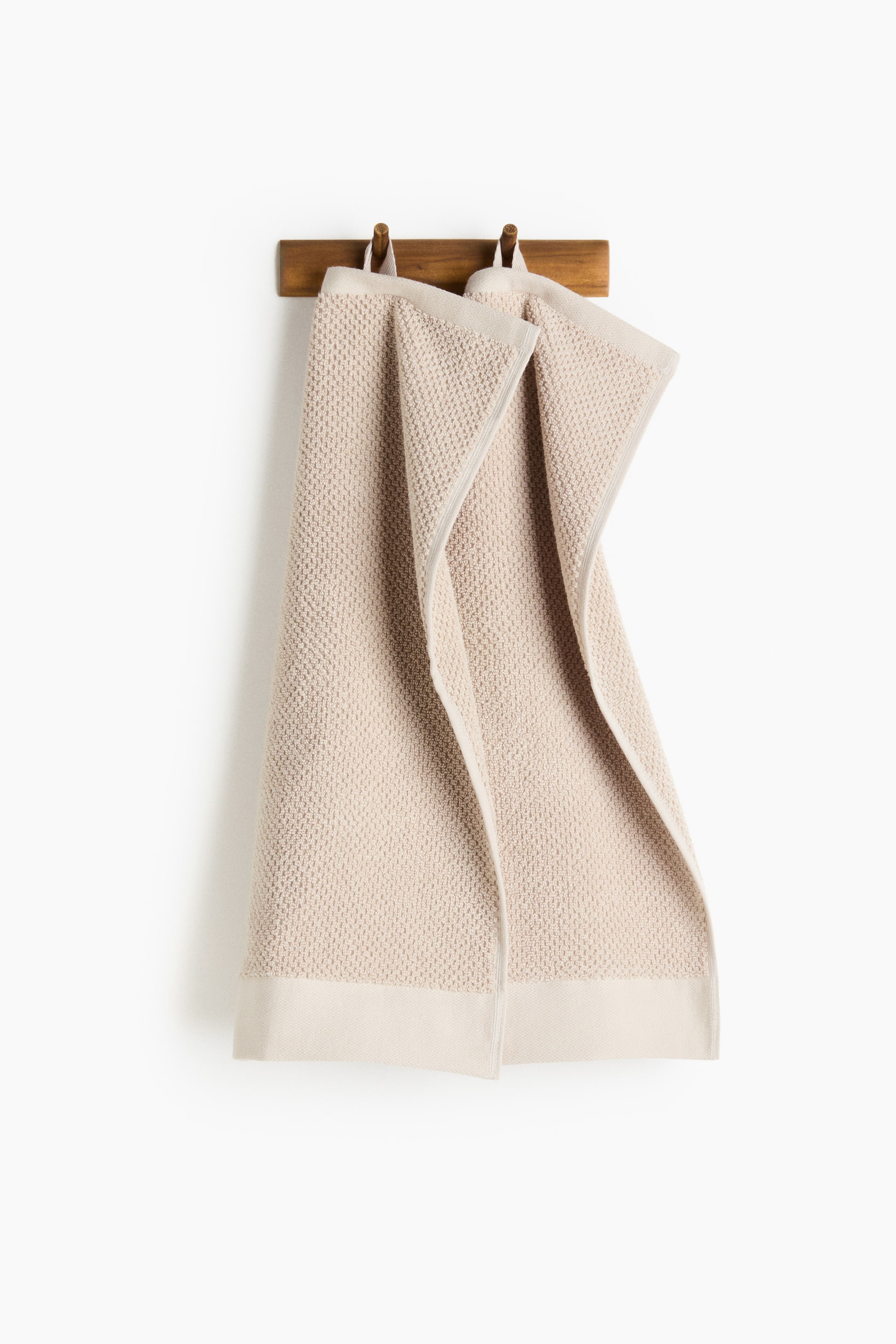 View larger image: Two light beige guest towels hang from a wooden rack. Each towel features a textured small square pattern, a smooth bottom border, and a visible loop at one short side for hanging.