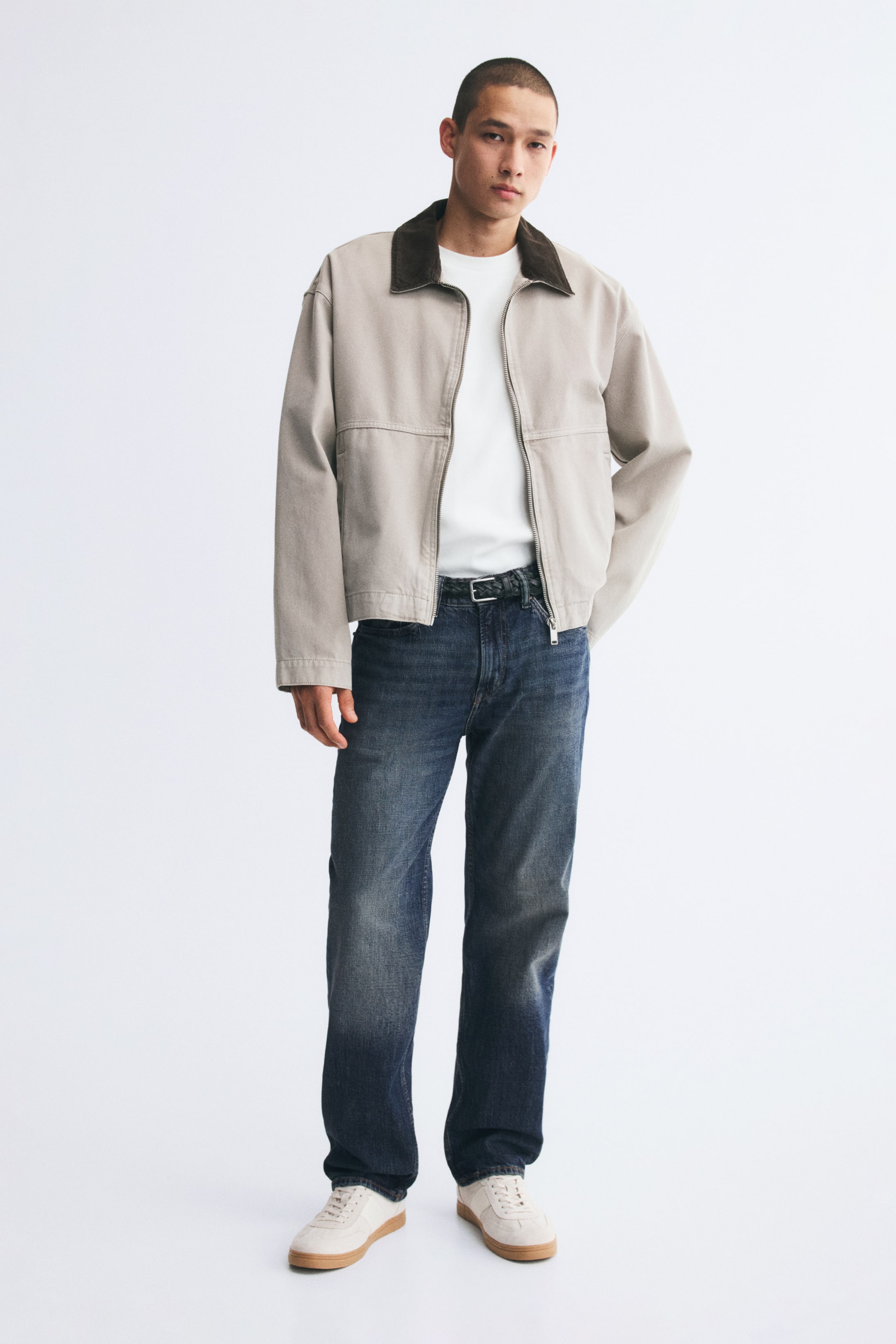 View larger image: Dark blue straight-leg denim jeans with subtle fading on the thighs are worn by the model. The jeans are styled with a braided black belt, a tucked-in white T-shirt, an open light-coloured jacket with a dark collar, and white sneakers.