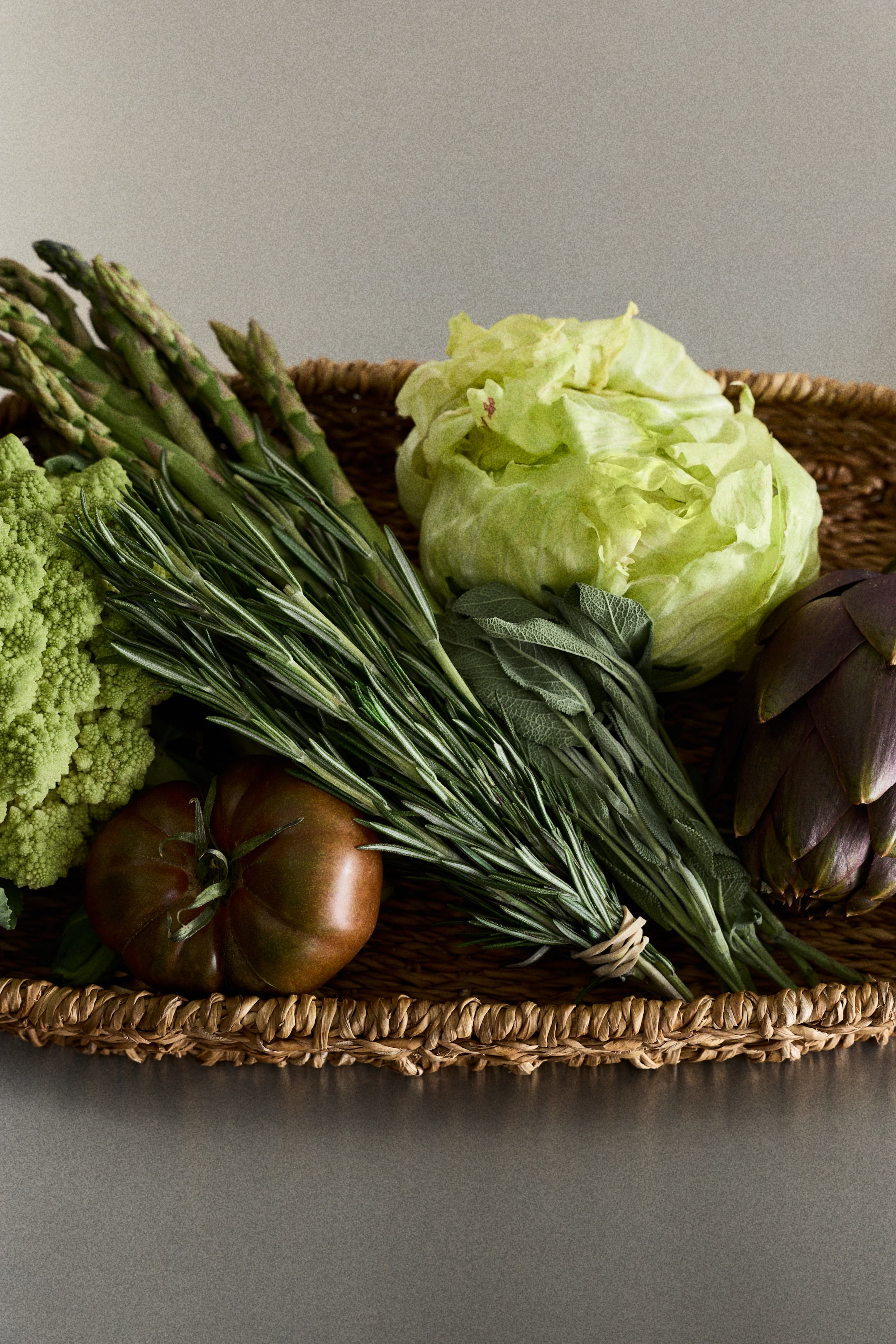 View larger image: A woven basket displays a collection of fresh produce, including green asparagus, light-green Romanesco broccoli, a head of light-green lettuce, a dark reddish-brown heirloom tomato, and a purple-green artichoke. Bundles of dark-green rosemary and gray-green sage are nestled among the vegetables.