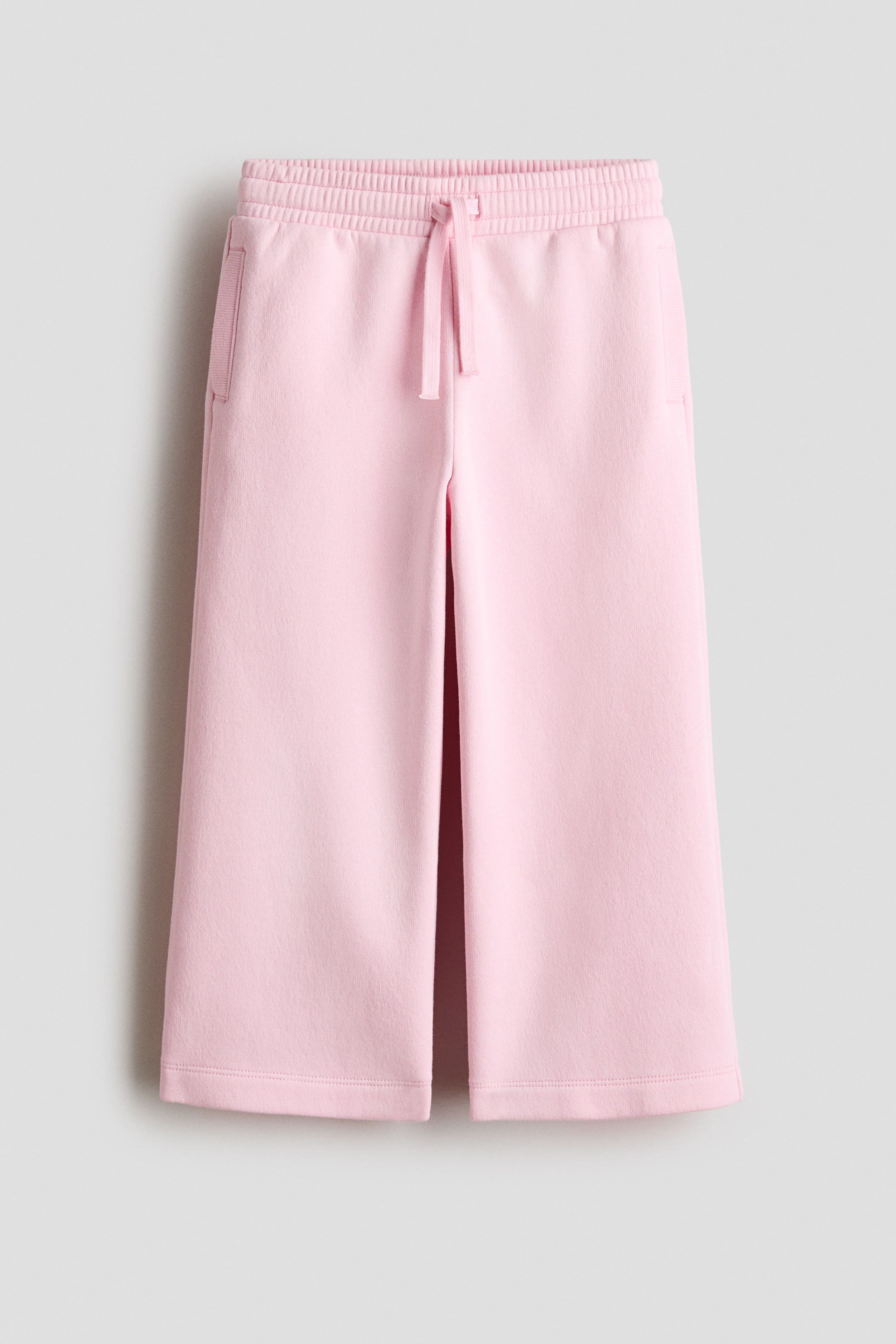 View larger image: Pink joggers are shown from the front, featuring an elasticated waist with a visible drawstring, side pockets, and wide legs. The fabric appears as a smooth, sweatshirt-like material.