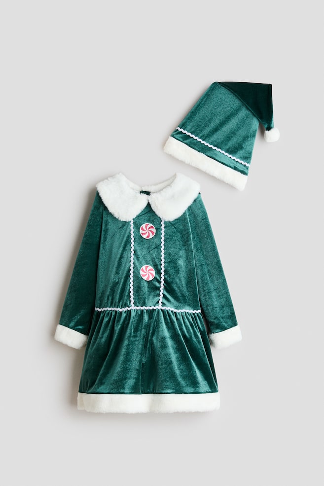 Ein grÃ¼nes Samtkleid und der dazu passende spitze Hut sind in einer Frontansicht zu sehen, mit weichem weiÃen Kunstpelzbesatz an Kragen, ÃrmelbÃ¼ndchen, Saum und Hutband. Das Kleid zeigt eine geraffte Taille, weiÃe Ric-Rac-Borte, die den mittleren Brustbereich einfasst, sowie zwei dekorative KnÃ¶pfe im Stil von rosa-weiÃen Pfefferminzbonbons. - 1