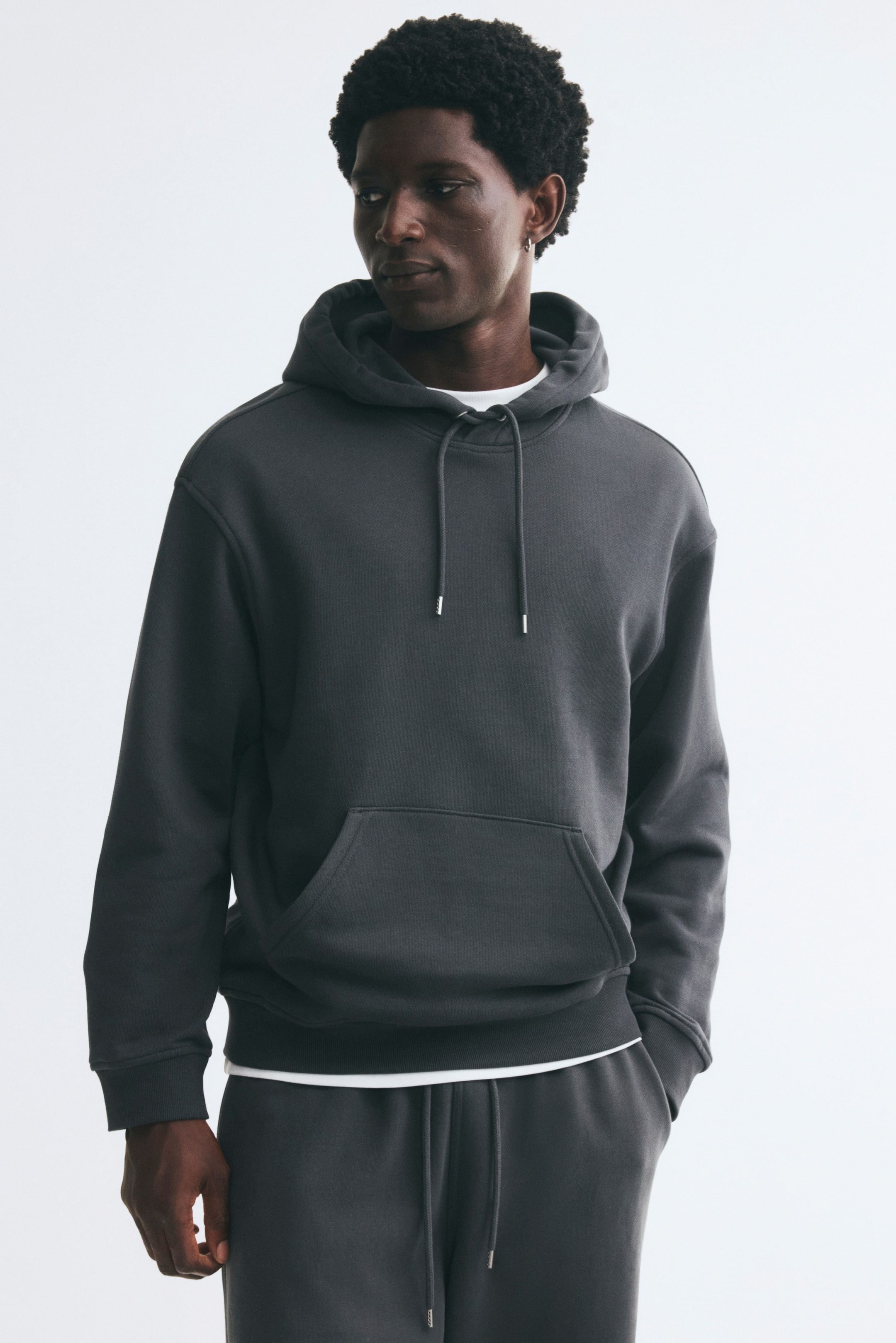 View larger image: A dark grey relaxed-fit hoodie is worn by the model, featuring a drawstring hood, dropped shoulders, a kangaroo pocket, and ribbed cuffs and hem. The hoodie is styled over a white T-shirt and paired with matching dark grey sweatpants.