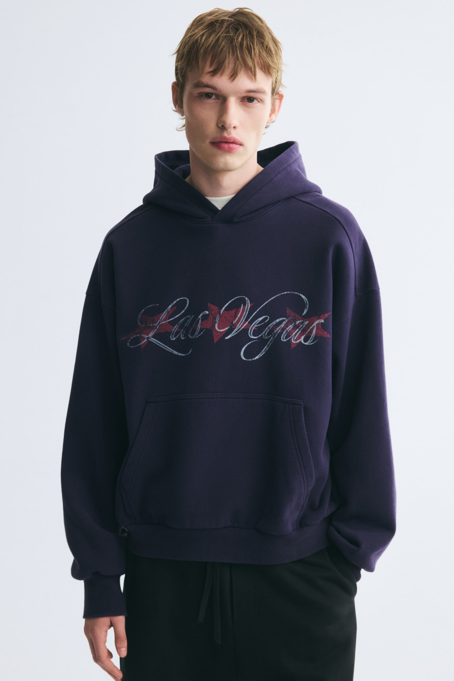 Má»t chiáº¿c hoodie oversized mÃ u tÃ­m Äáº­m, ná»i báº­t vá»i há»a tiáº¿t chá»¯ "Las Vegas" viáº¿t tay cÃ¡ch Äiá»u á» ngang ngá»±c, Äang ÄÆ°á»£c ngÆ°á»i máº«u máº·c. Hoodie cÃ³ vai xuÃ´i, tÃºi kangaroo vÃ  bo tay bo gÃ¢n, ÄÆ°á»£c phá»i cÃ¹ng quáº§n dÃ¢y rÃºt mÃ u Äen. - 1